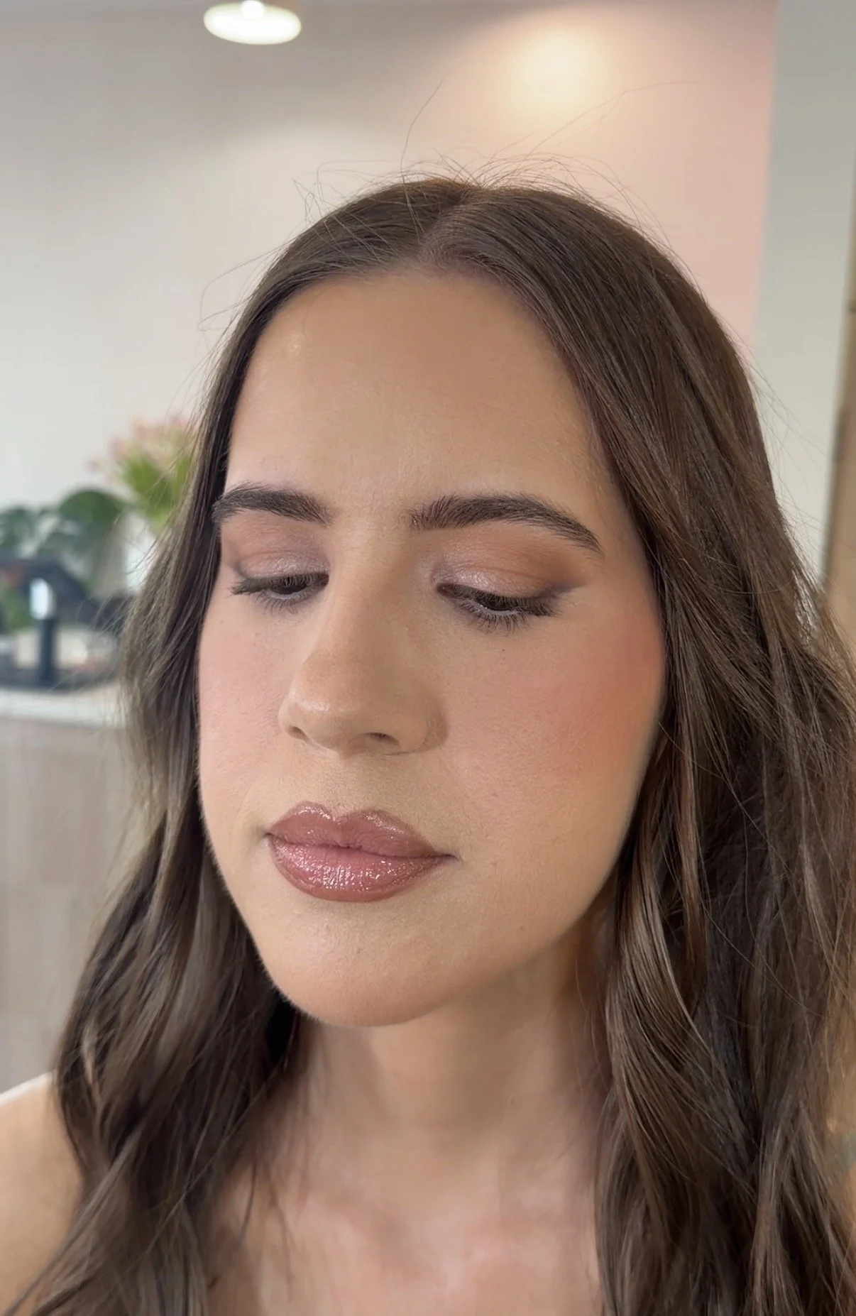 Close-up of a woman with long, wavy brown hair and natural makeup, looking slightly downward with closed eyes, in a bright indoor setting.