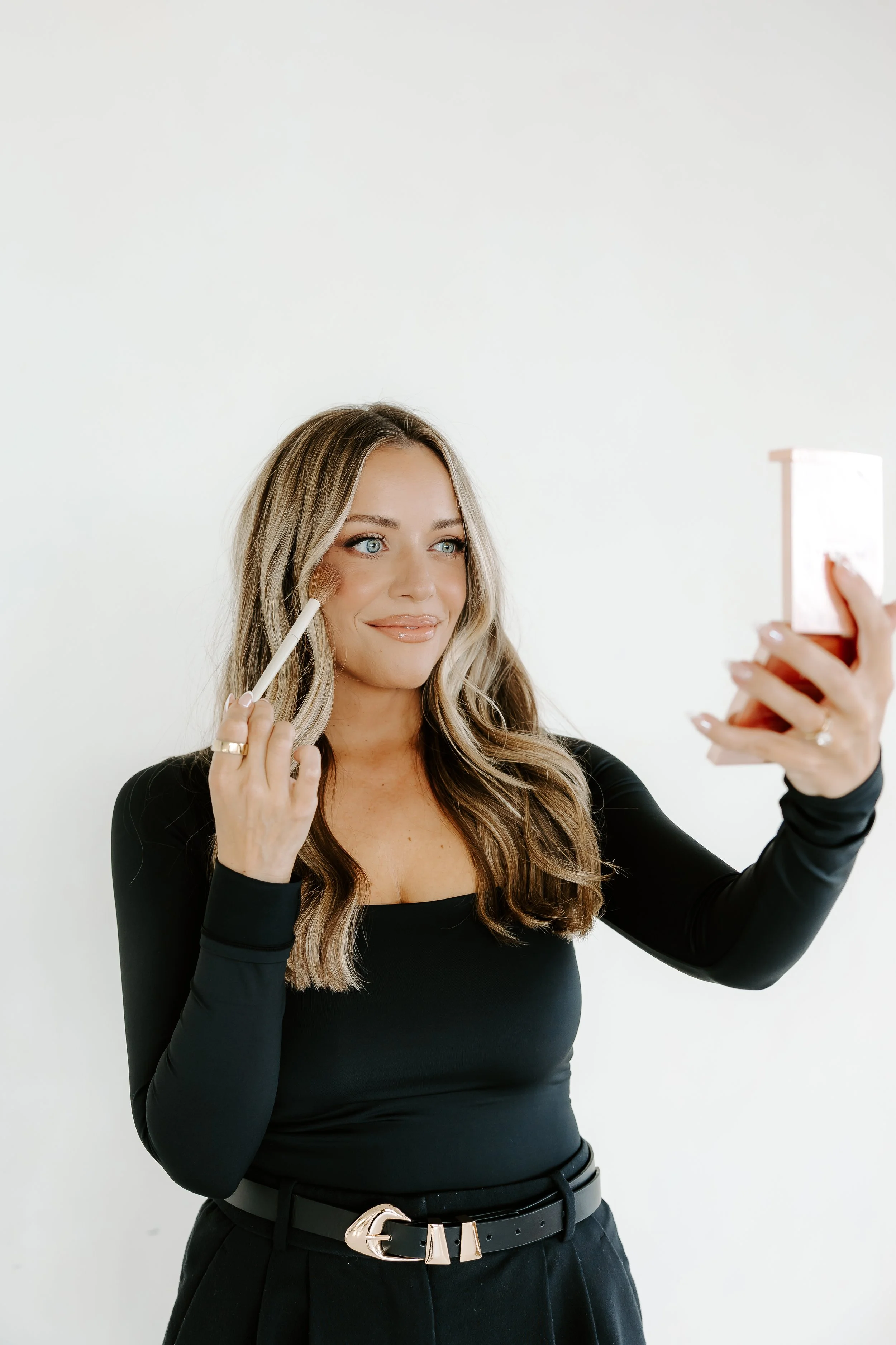 A woman with long wavy blonde hair and blue eyes is smiling while applying makeup with a brush in her right hand, and she is taking a selfie with her left hand holding a smartphone. She is wearing a black long-sleeve top and high-waisted black pants with a gold belt, standing against a plain white background.
