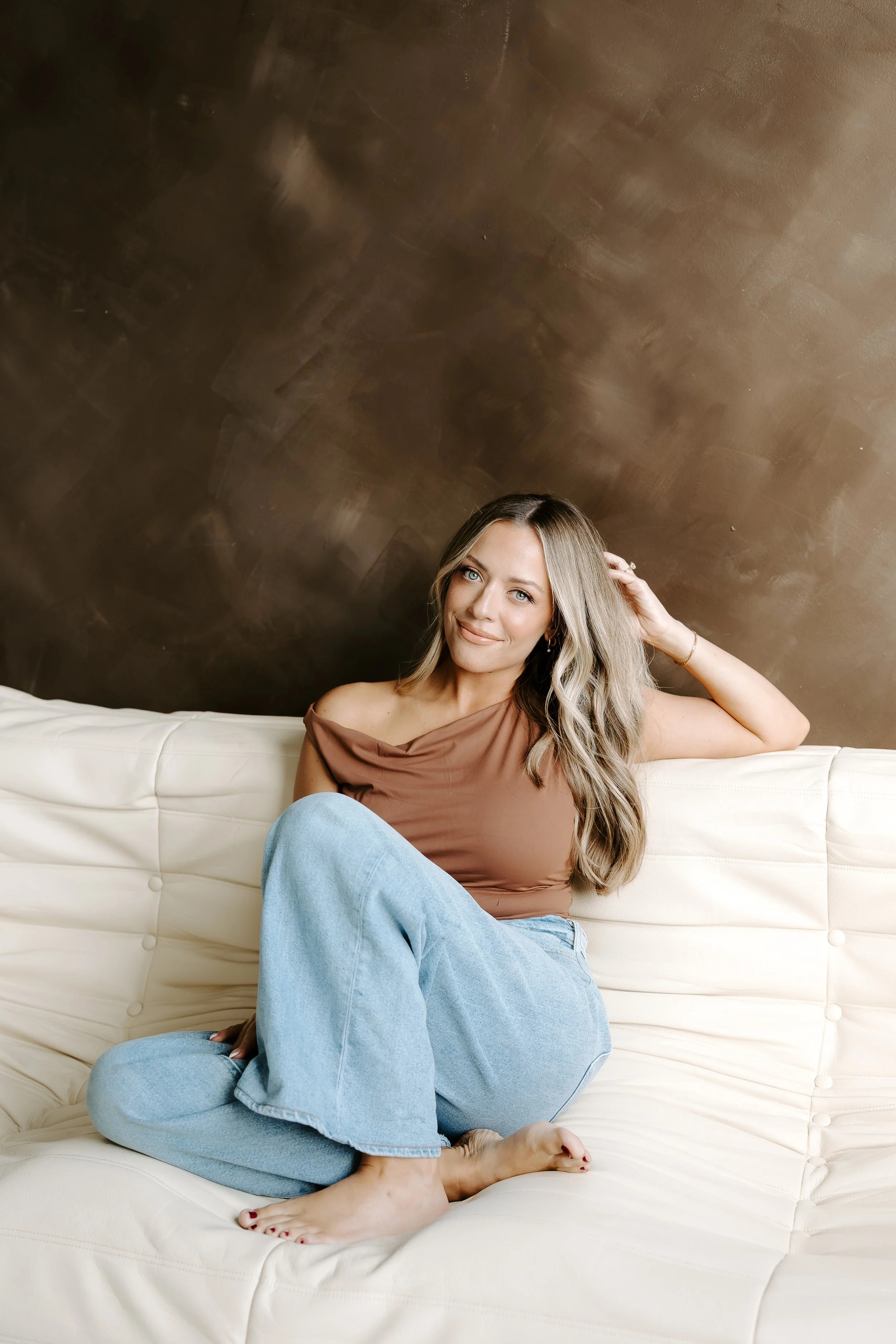 A woman with long blonde hair, blue eyes, wearing a brown off-the-shoulder top and light blue jeans, sitting on a white tufted couch against a brown wall. She is smiling, resting her head on her hand, with one leg crossed over the other, barefoot with painted toenails.