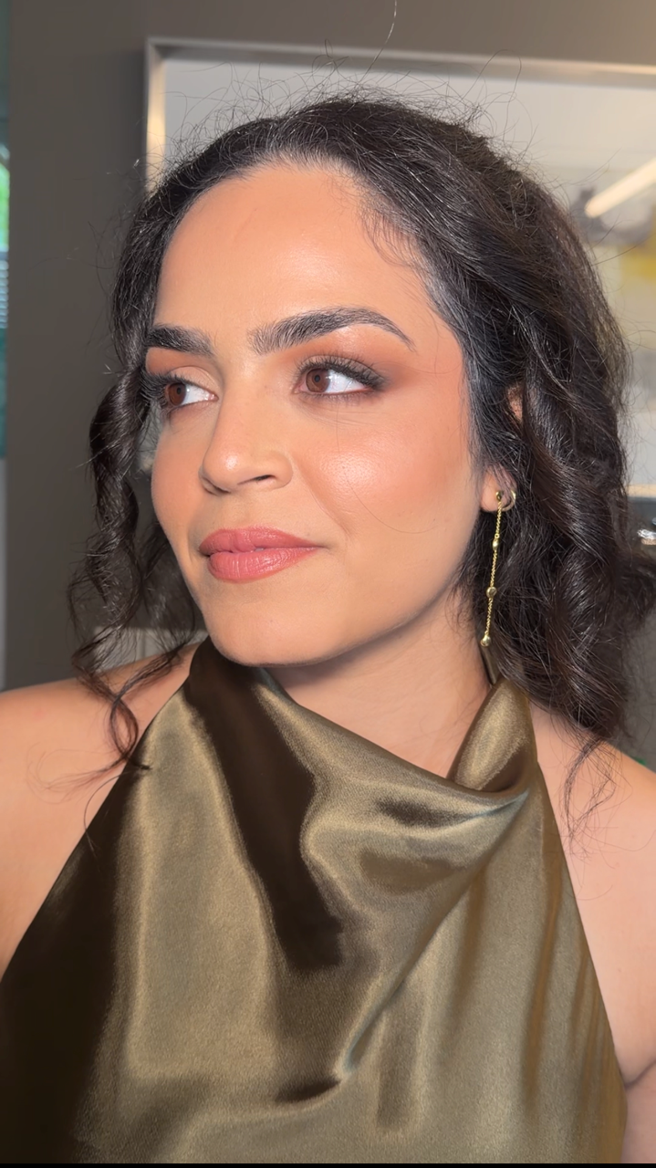 A woman with dark, curly hair wears a shiny, olive-green satin dress and a long, gold dangling earring, posing indoors with a neutral expression.