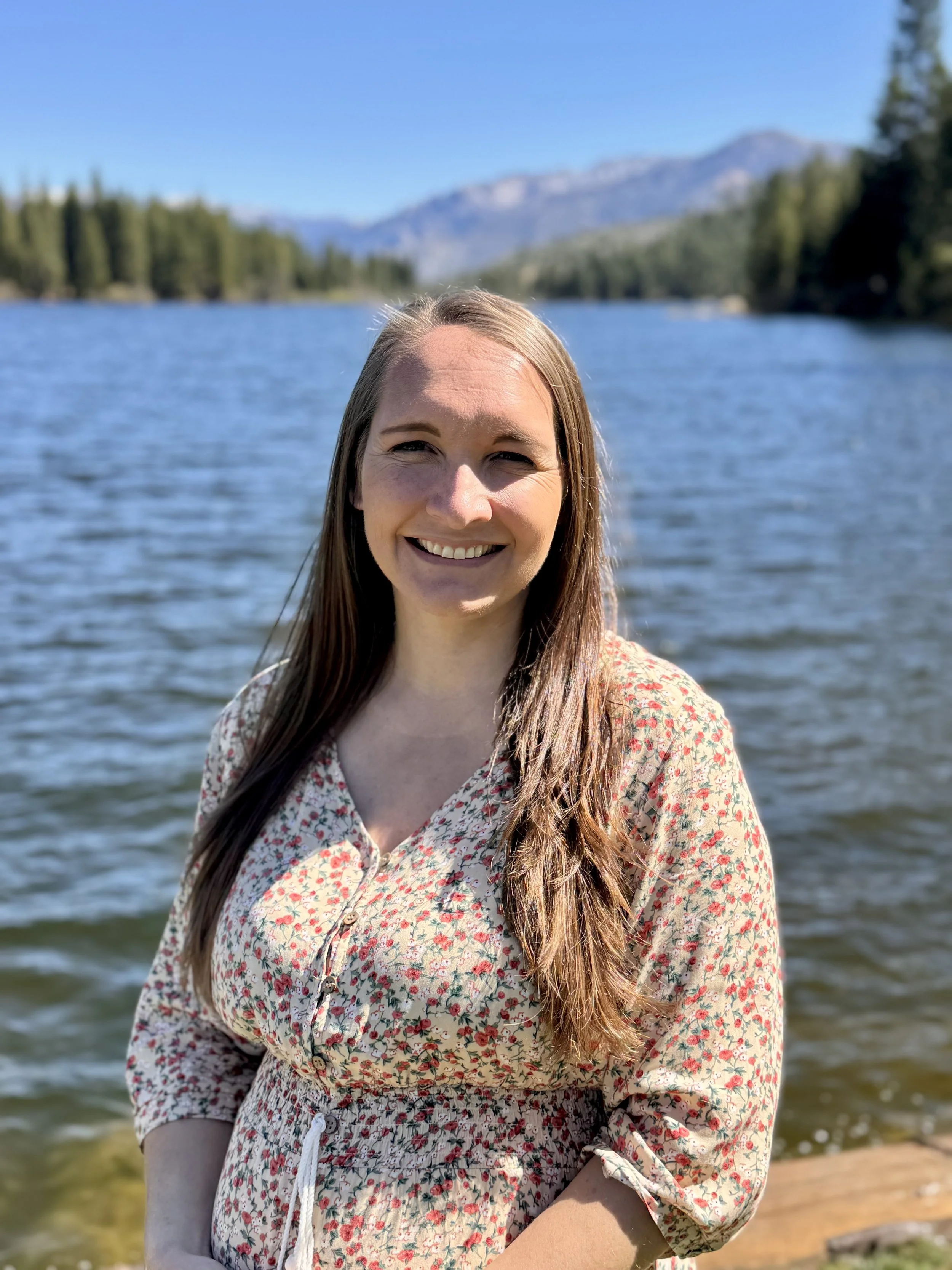 A smiling woman with long brown hair standing near a body of water with mountains in the background on a clear, sunny day.