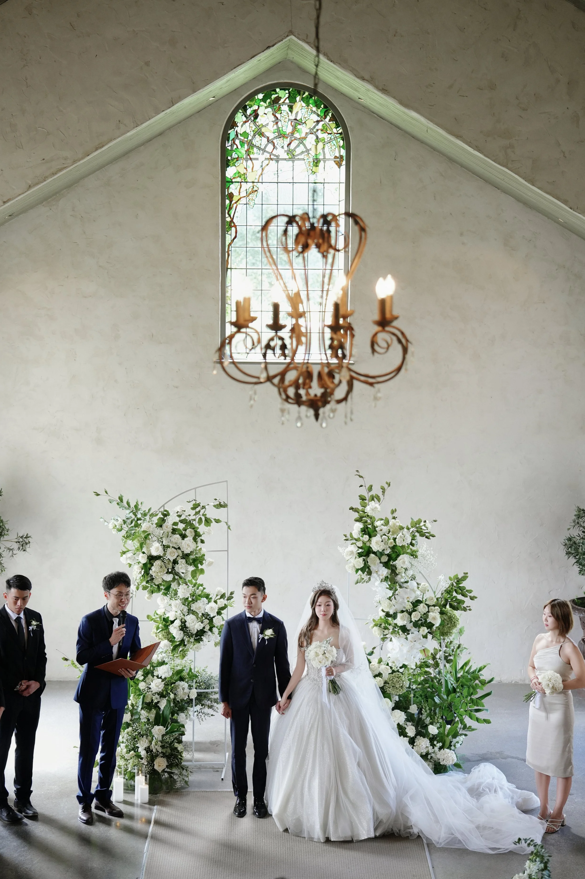 A wedding ceremony taking place in a church with a bride and groom holding hands, surrounded by floral arrangements, with officiant and bridesmaid present, under a chandelier and stained glass window.