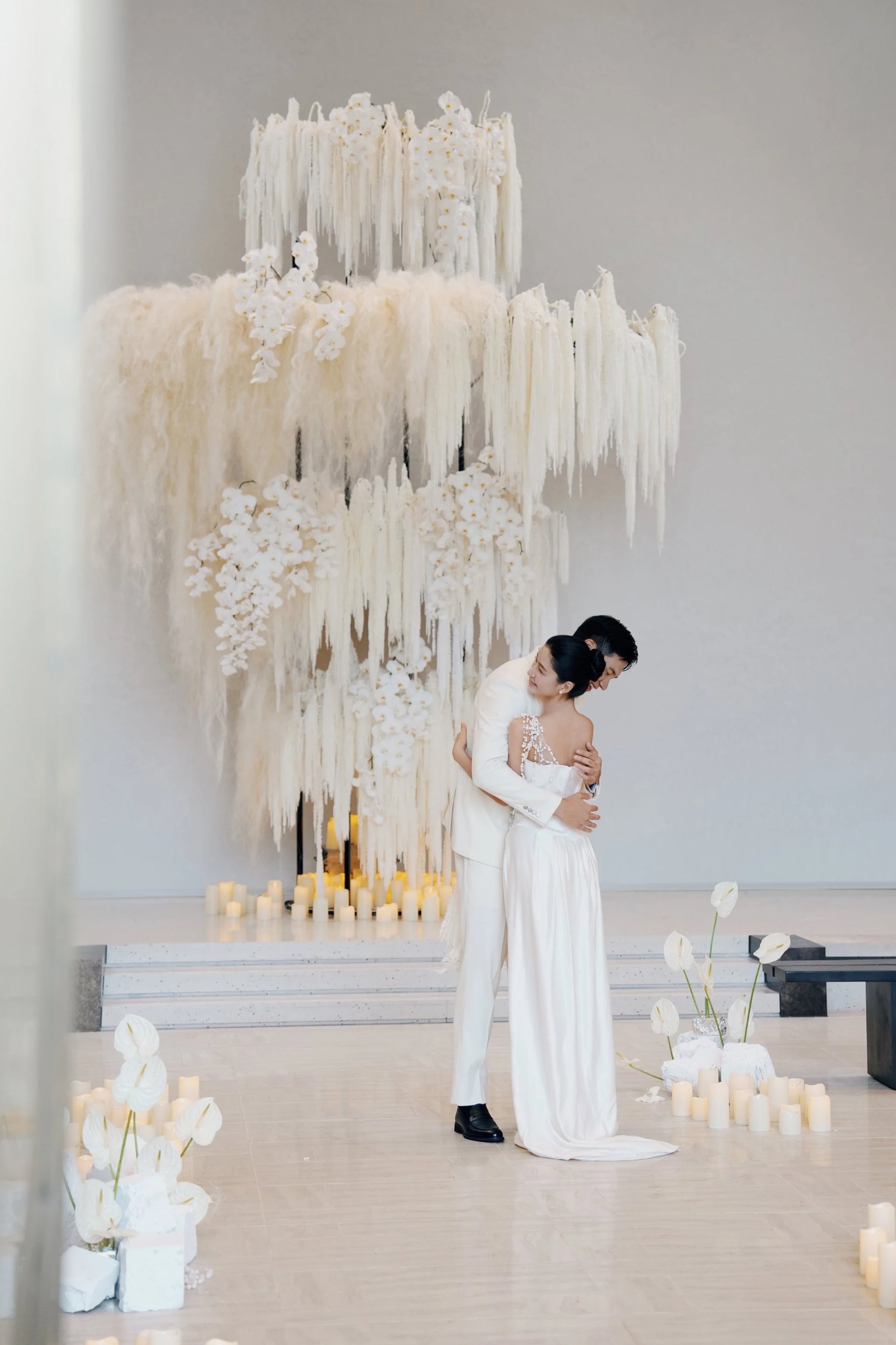 A bride and groom embracing during a wedding ceremony in front of a large, white, abstract floral art installation with candles and white flowers on the floor.