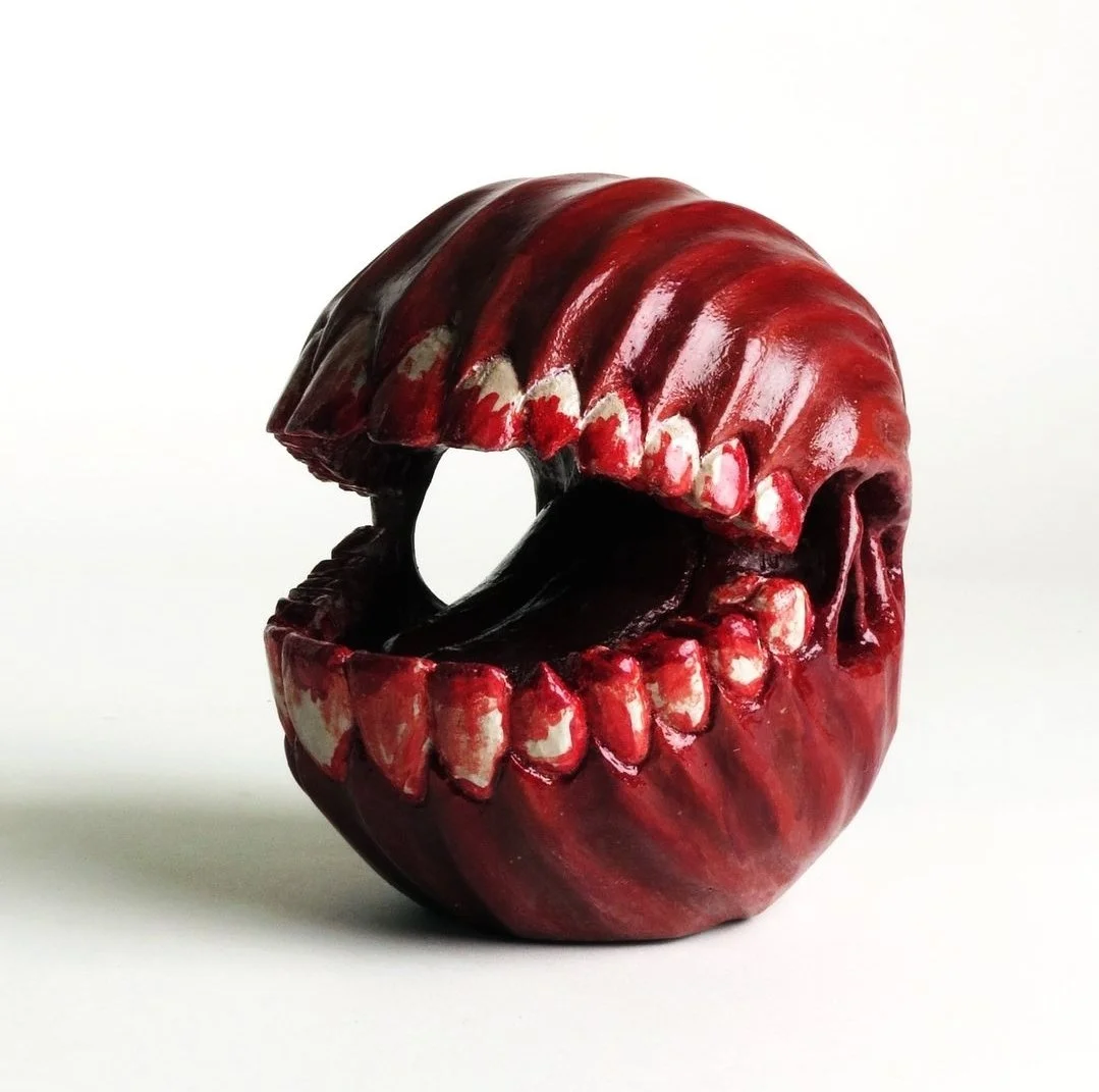 Sculpture en forme de tête de crâne avec une bouche ouverte, couleur rouge foncé avec des détails en blanc.