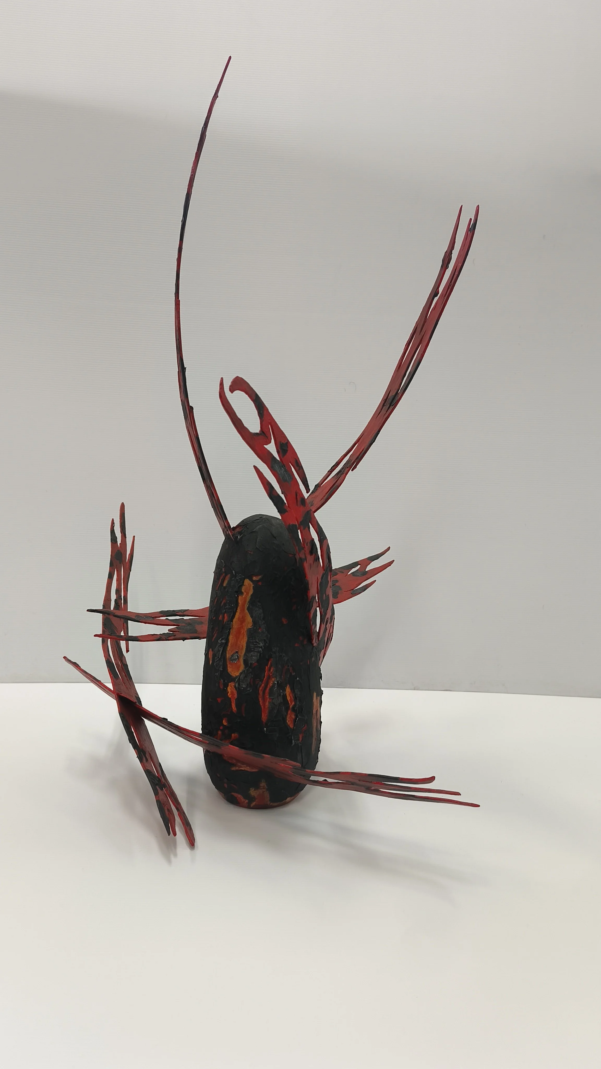 Une sculpture artistique avec une base noire et rouge, avec des éléments en forme d'antennes ou de branches rouges et noires, disposée sur une surface blanche devant un mur gris.