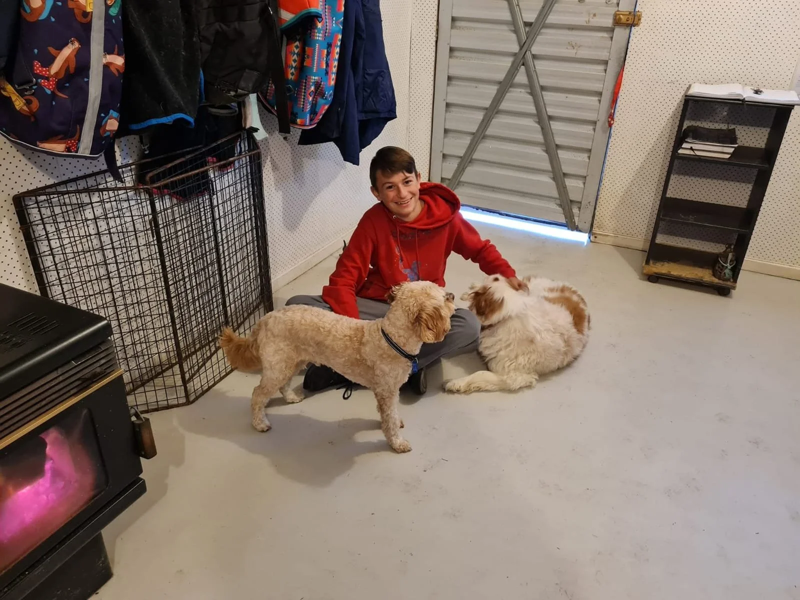 A smiling boy in a red hoodie sitting on the floor with two dogs, one tan and one white, inside a garage or storage area with various backpacks hanging on the wall and a metal gate.