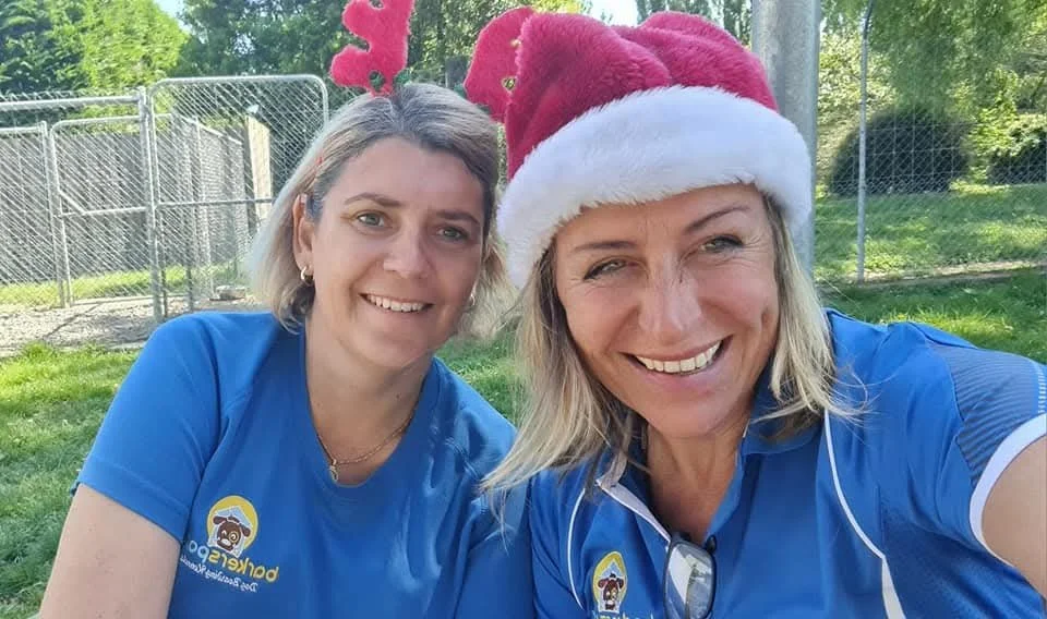 Two women taking a selfie outdoors, smiling, with a fence and trees in the background. One woman wears a blue shirt and a Santa hat, the other wears a blue shirt and pink reindeer antlers.