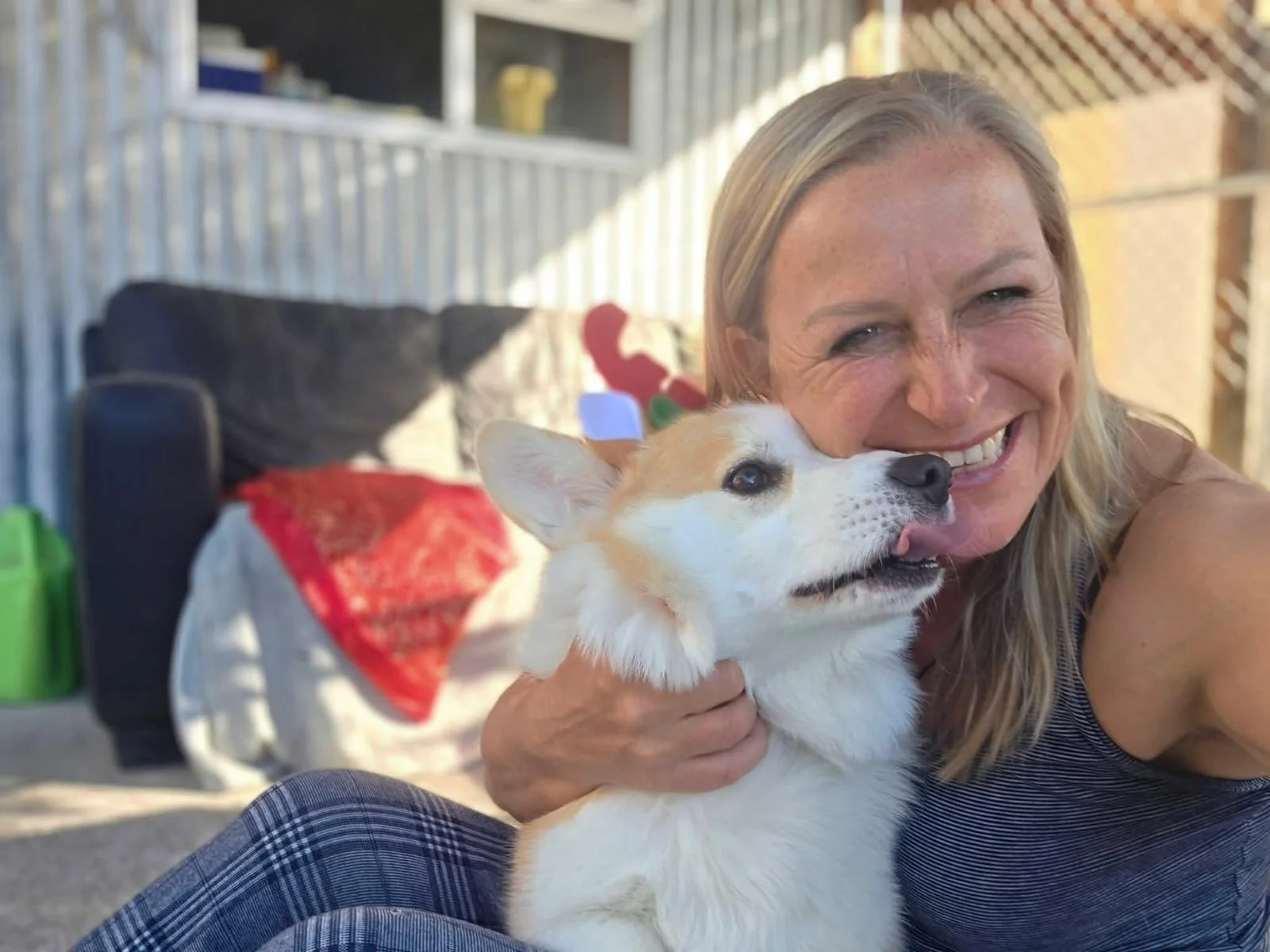 A woman with blonde hair smiling and holding a cute corgi puppy outside on a sunny day.