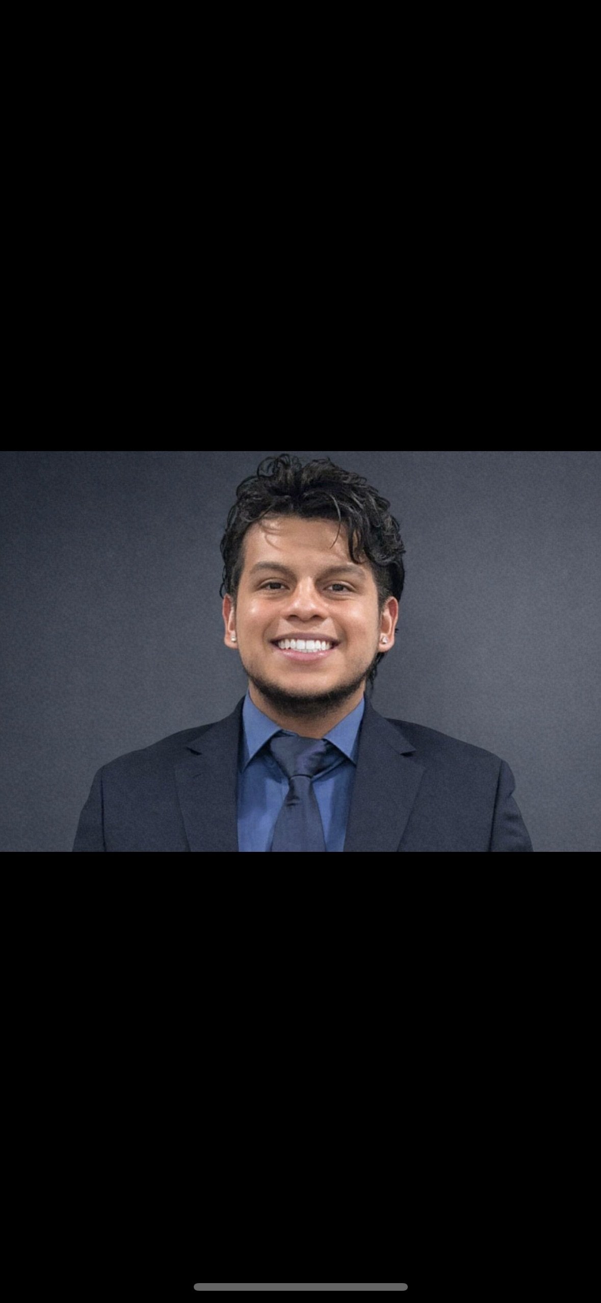 Portrait of a young man in a dark suit, blue dress shirt, and matching tie, smiling against a dark background.