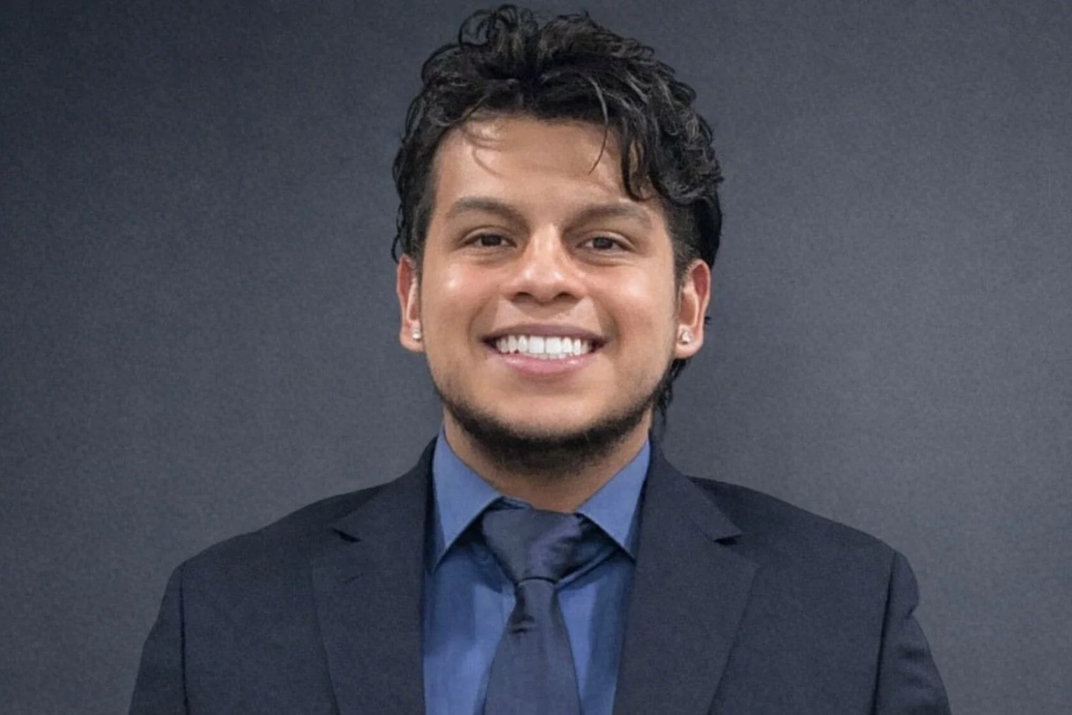 A young man dressed in a dark suit and blue shirt, smiling at the camera, with a dark grey background.
