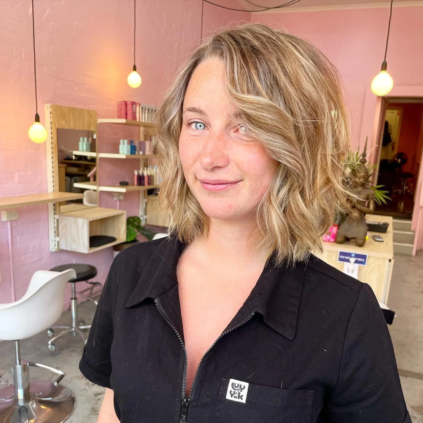 A woman with wavy blonde hair and blue eyes smiles in a hair salon with pink walls and modern decor.