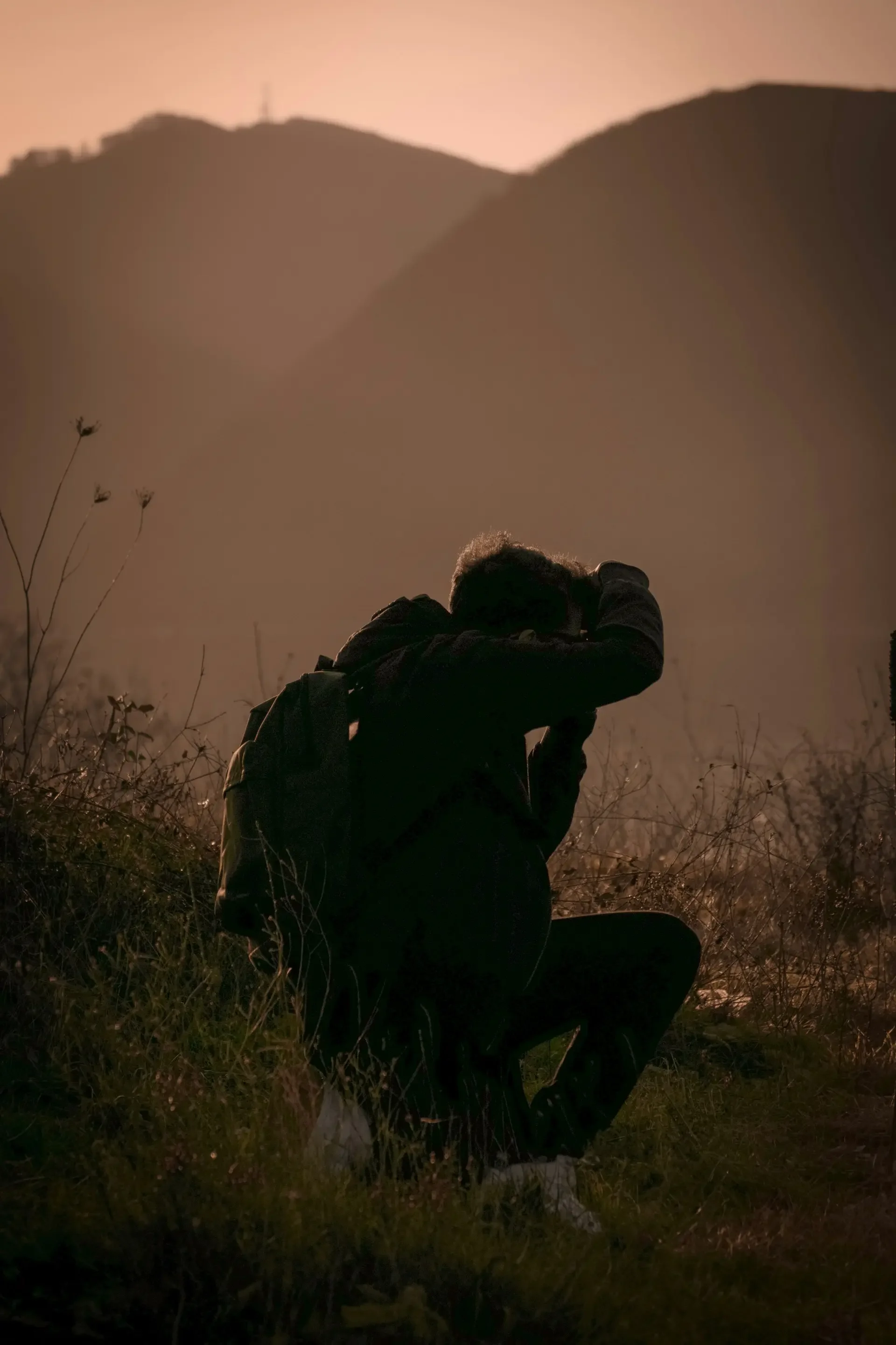 A person sitting on the ground holding a camera, with mountains in the background and a soft pinkish sky.