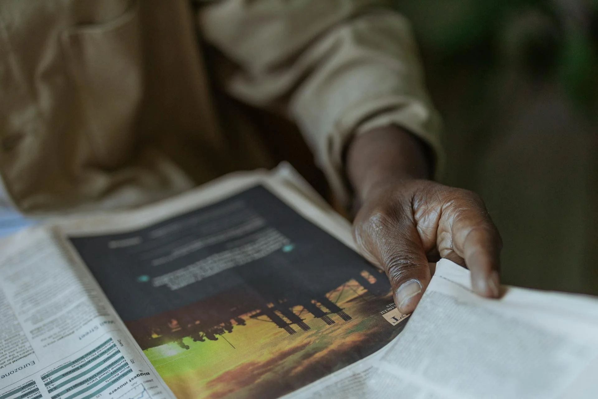 Close-up of a person turning the pages of a newspaper on a table, showing part of a landscape image and text.