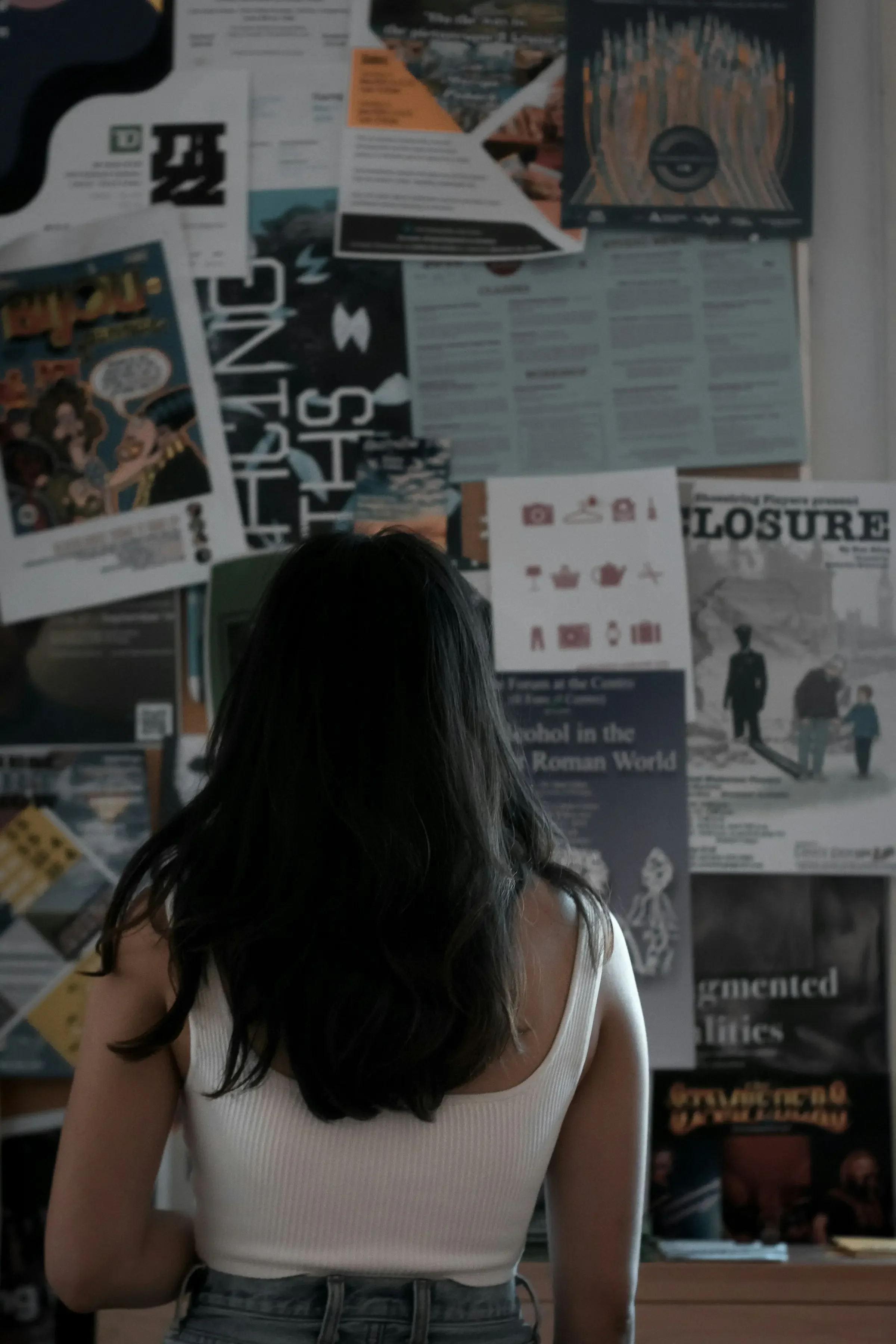 A woman with long dark hair wearing a white tank top and gray jeans standing in front of a bulletin board covered with various posters and papers.
