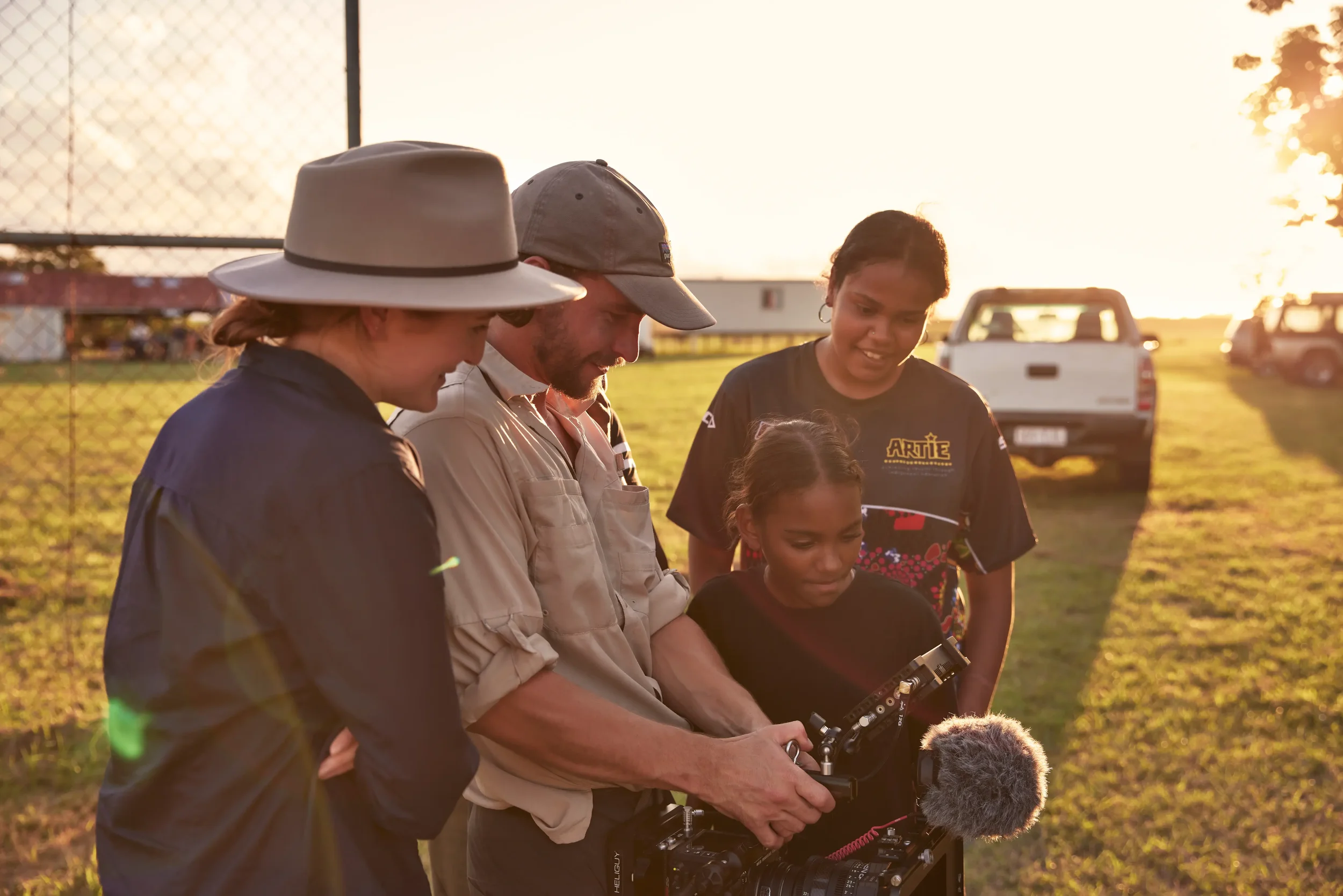 A group of four people, including children and adults, gathered outdoors at sunset, looking at the screen of a video camera, with a background of a chain-link fence, parked trucks, and a grassy field.