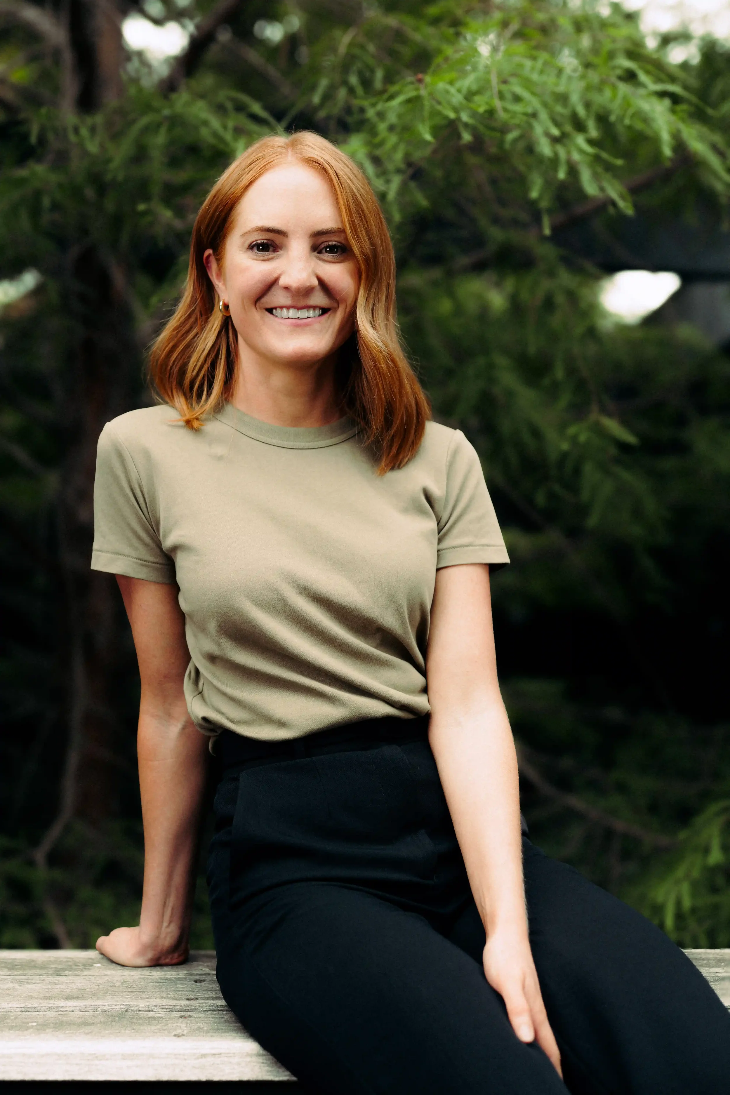 A woman with shoulder-length red hair, wearing a green t-shirt and black pants, sitting outdoors on a wooden surface, smiling, with green trees and foliage in the background.