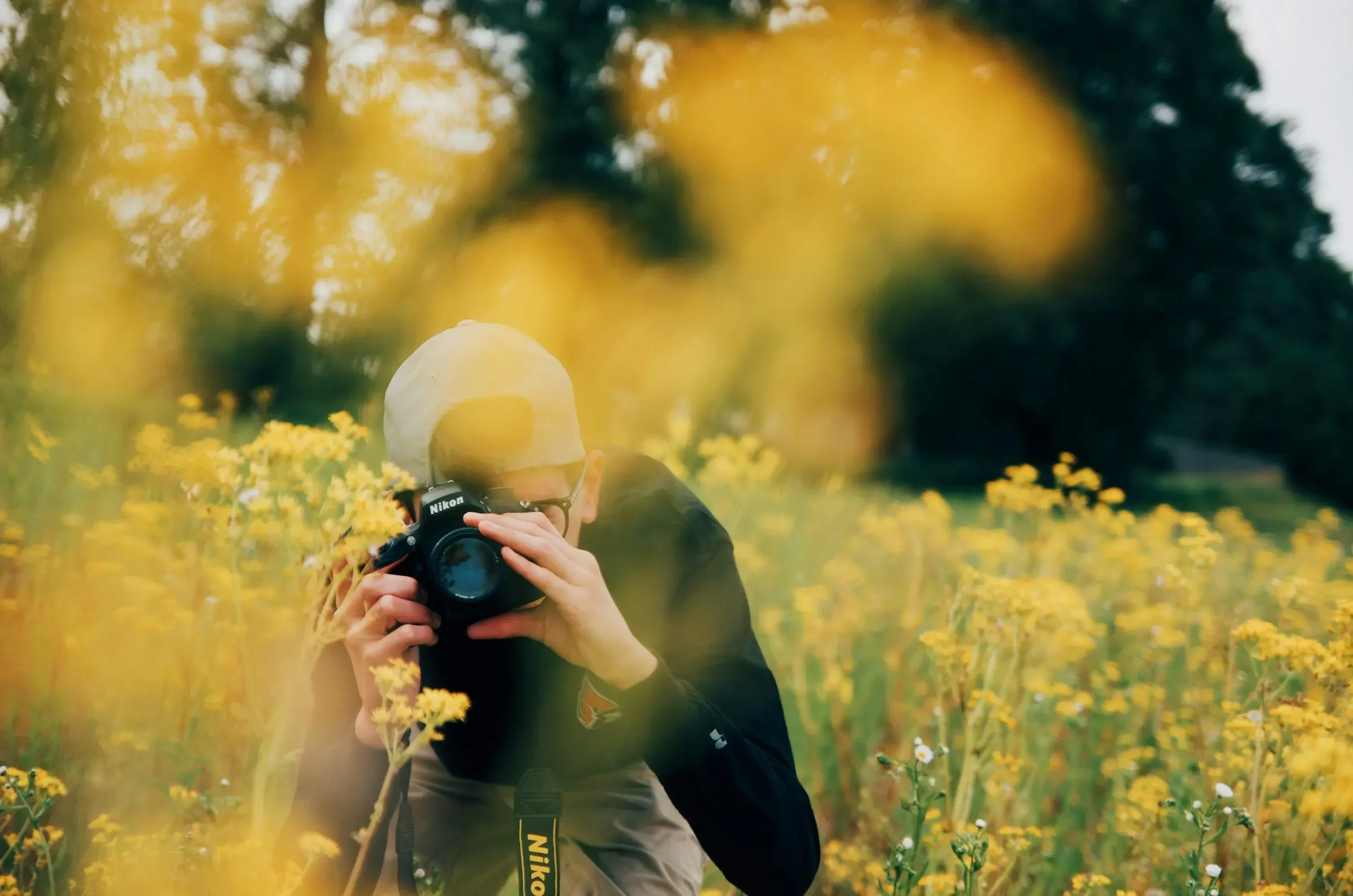 Person wearing a white cap and glasses holding a Nikon camera, surrounded by yellow flowers in a field with blurred trees and bright light in the background.