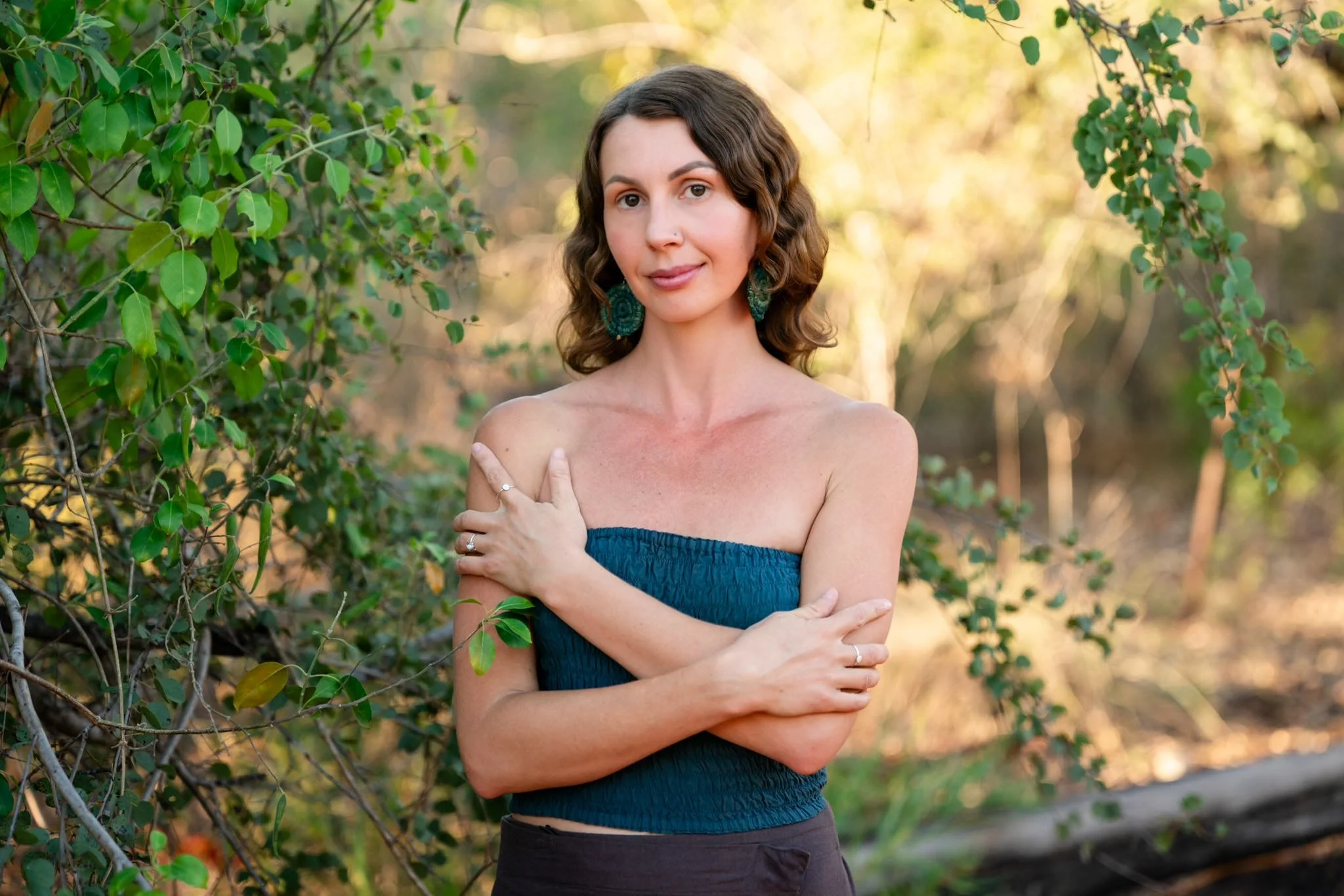 A woman with wavy brown hair and large teal earrings stands outdoors in front of green foliage, wearing a strapless blue top and has her arms crossed over her chest.