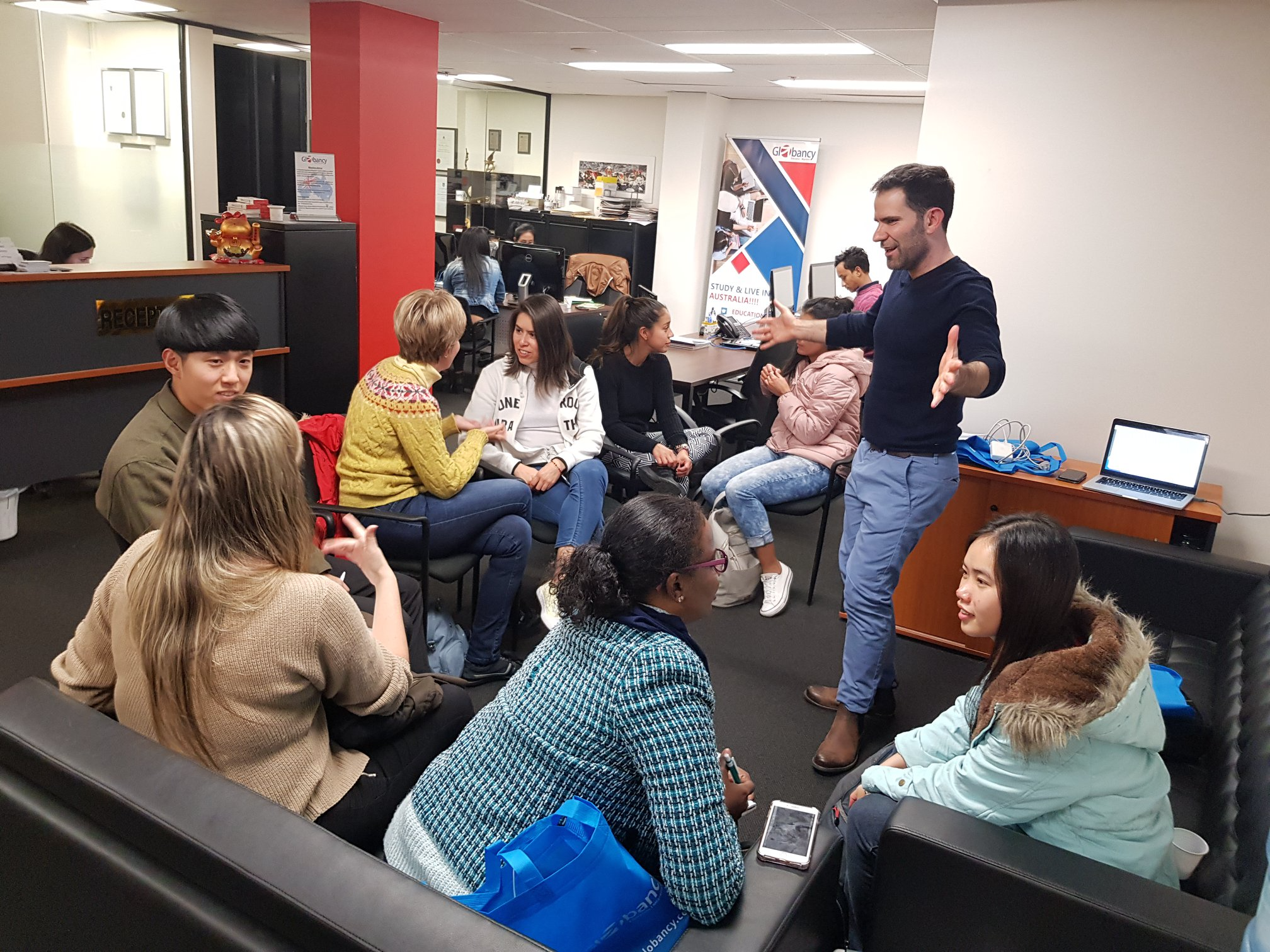 A group of diverse people are sitting in a waiting area, engaged in conversation and using their phones. A man stands in front of them, speaking or presenting. In the background, there are desks with computers and office supplies, and a large banner 