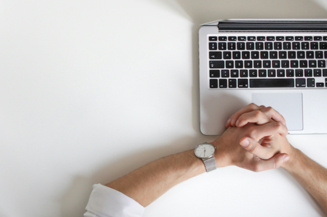 Person with a watch on their left wrist sitting at a white desk with a laptop, hands clasped near the laptop.