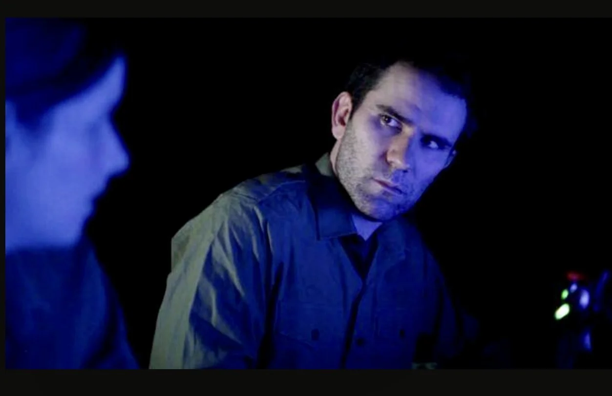 A man looking toward a woman dark room illuminated by blue light, engaging in a serious conversation.