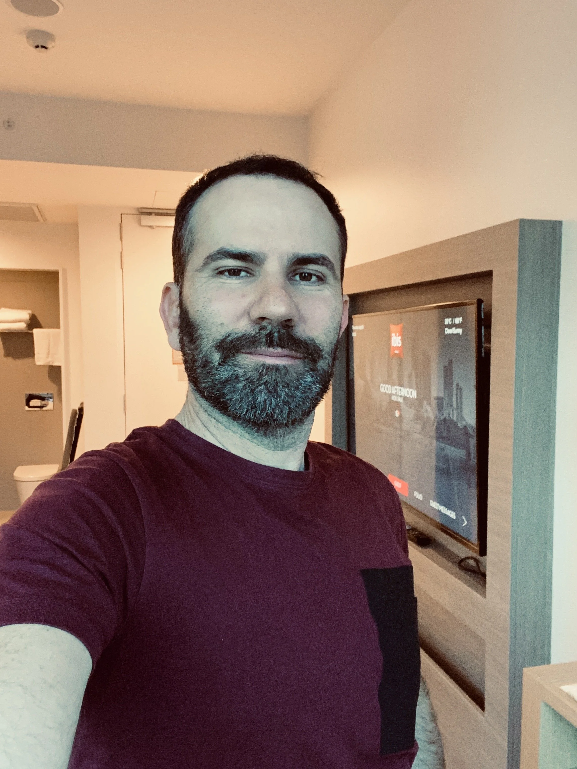 A man taking a selfie indoors, wearing a maroon shirt with long beige sleeves, with a television screen behind him displaying various content, in a room with beige walls and a standing heater.