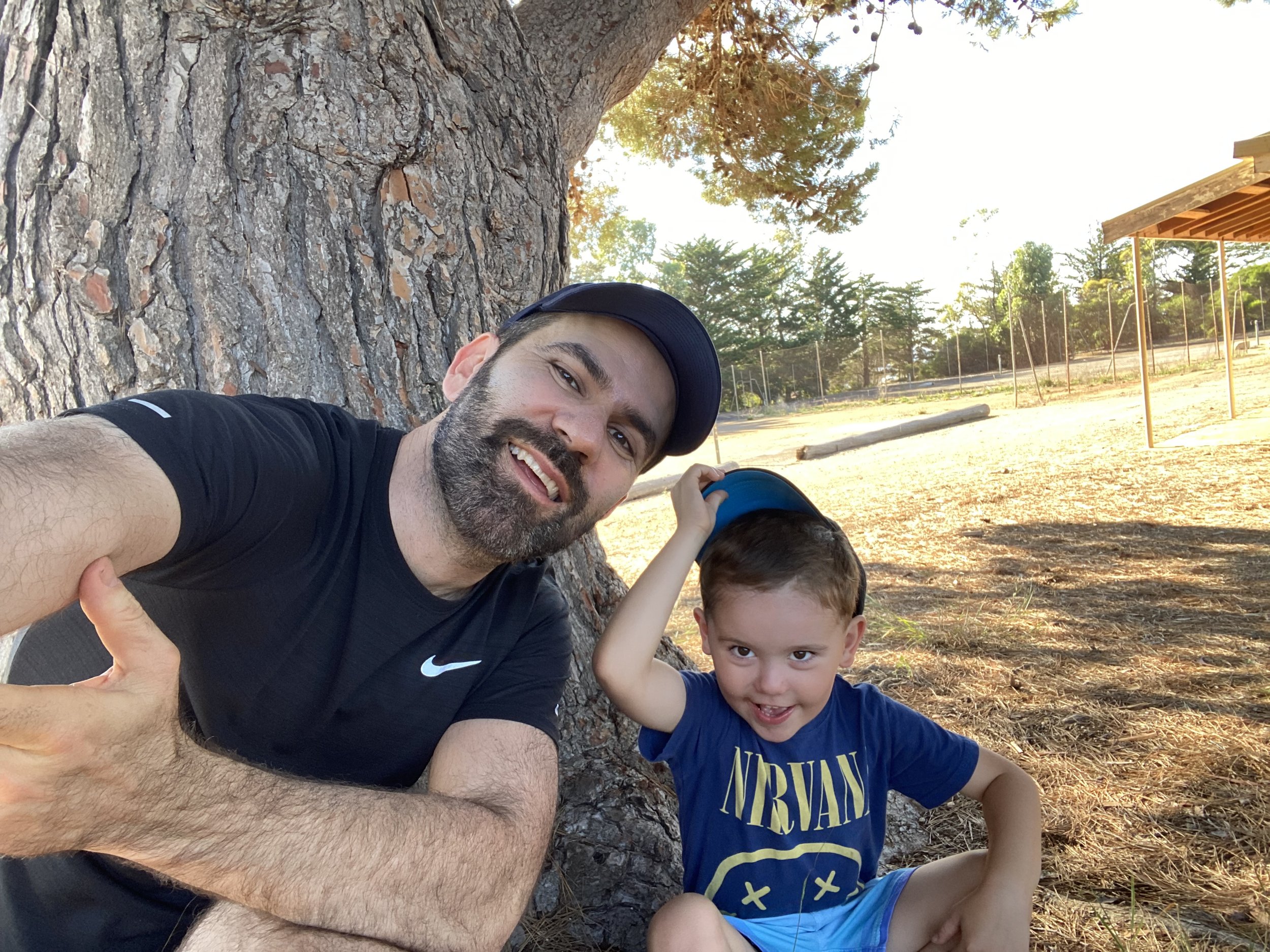 A man and a young boy taking a selfie next to a large tree trunk outdoors on a sunny day. The man is smiling and wearing a black shirt and a cap, and the boy is smiling and wearing a blue Nirvana t-shirt. The background shows trees and a park or spor