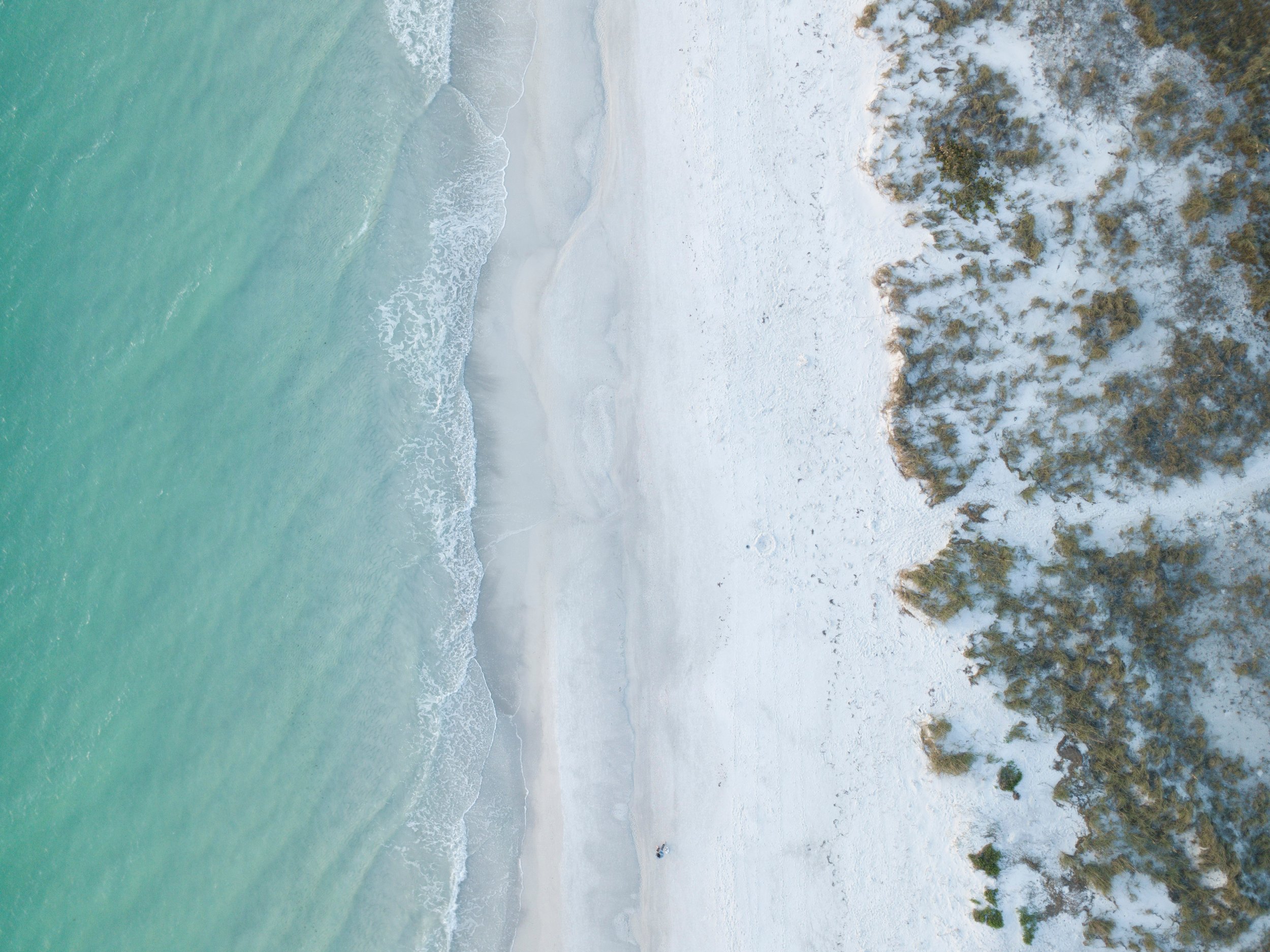 An aerial view of a beach with clear turquoise water, white sand, and patches of vegetation near the shore.