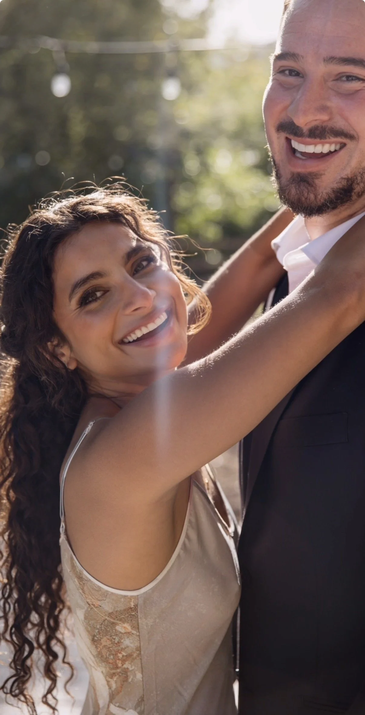 Bride with soft glam makeup and long curly hair smiling while embracing groom during an outdoor wedding