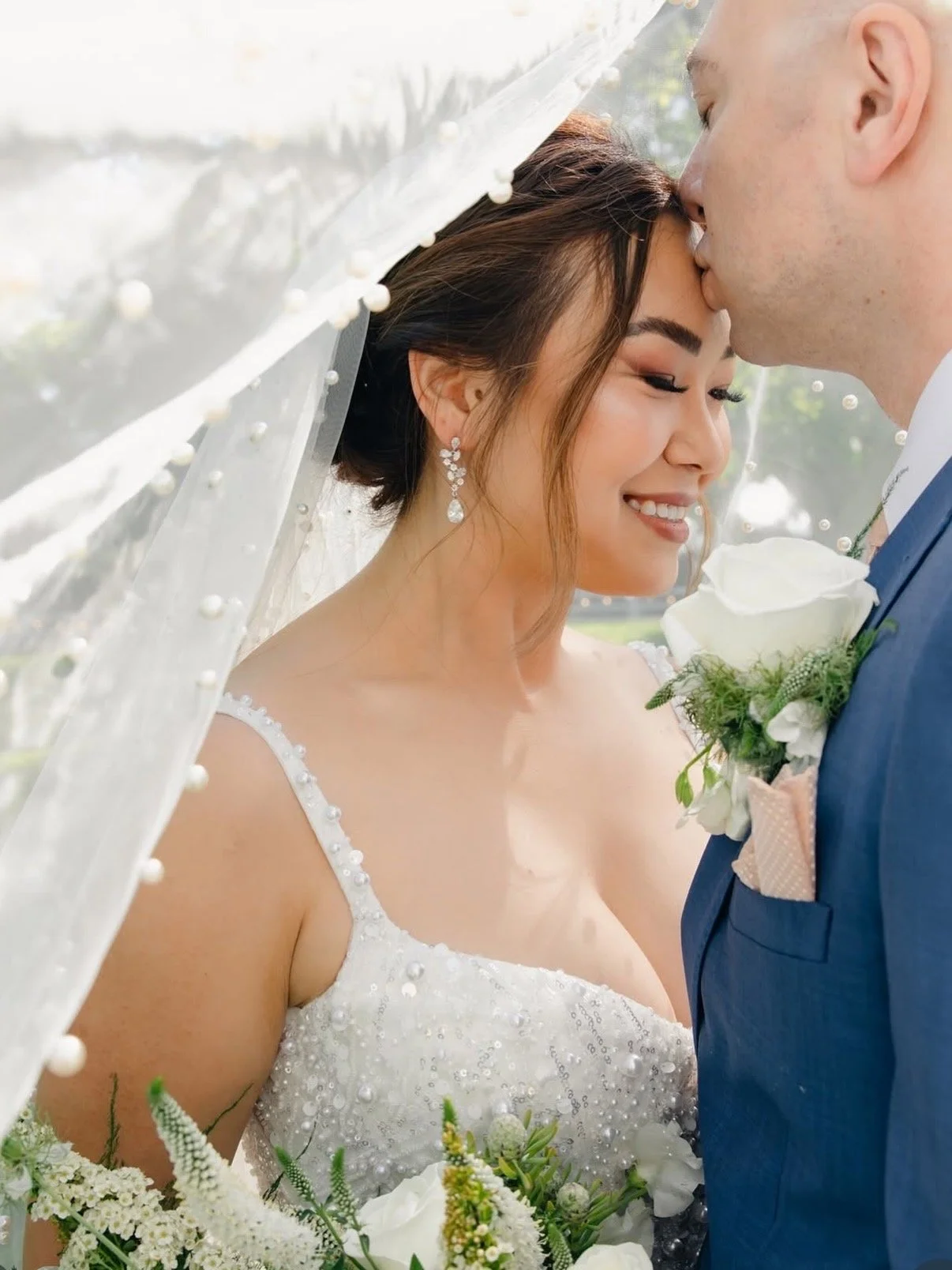 Close-up of a smiling bride wearing a delicate pearl veil and white floral dress, showcasing a soft glam makeup look.