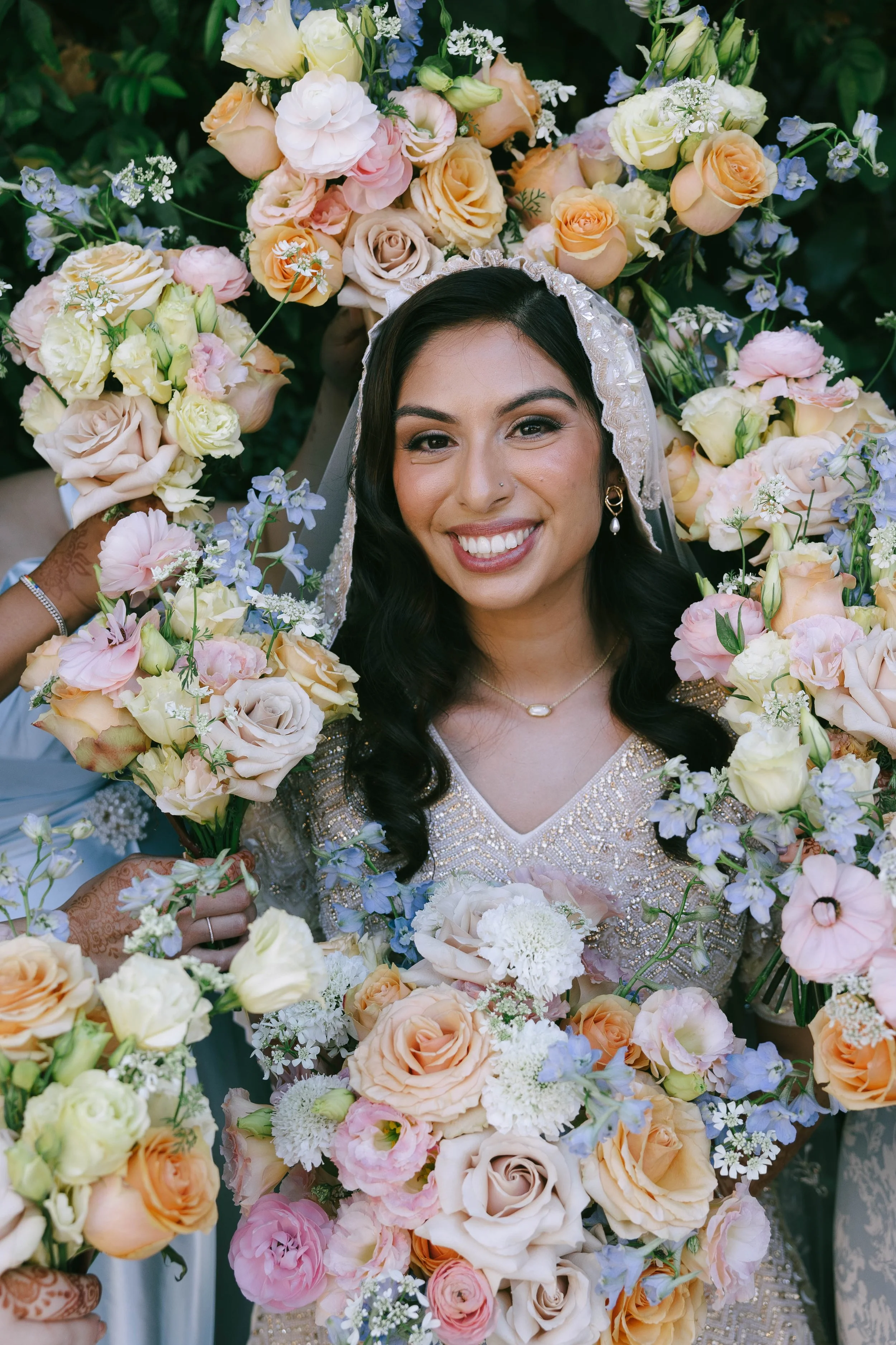 A smiling woman at a wedding surrounded by a large bouquet of pastel-colored roses, lisianthus, and other flowers.