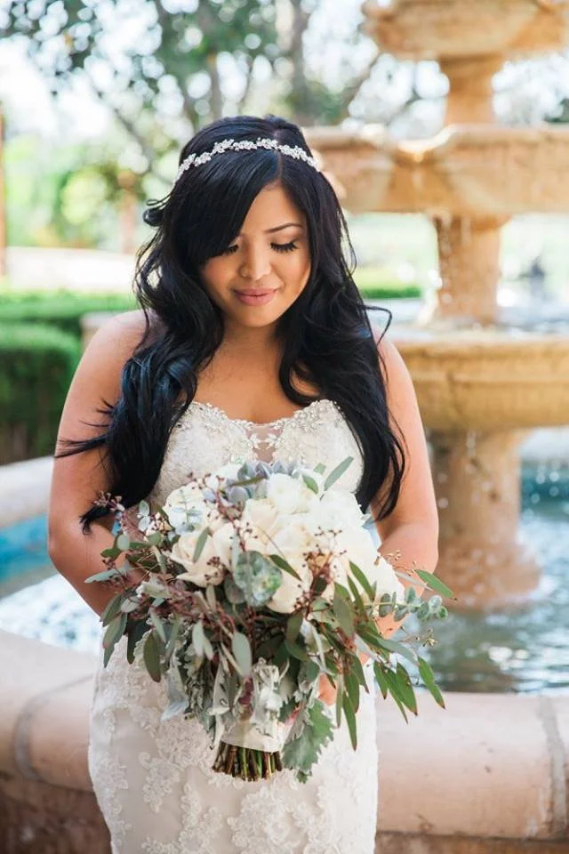 Bride with soft glam makeup and long loose waves holding a bouquet outdoors by a fountain.