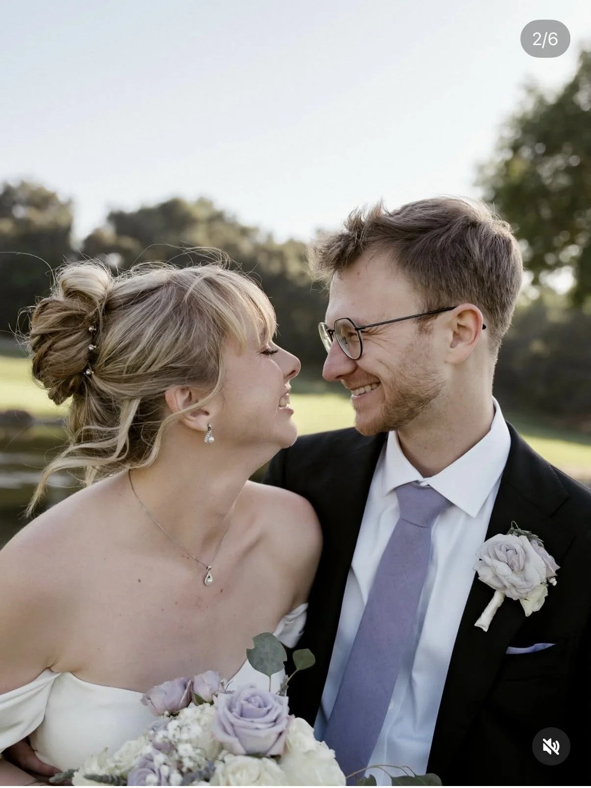 A newlywed couple dressed in wedding attire smiling and leaning close to each other outdoors on a sunny day, with green trees and a bright sky in the background.