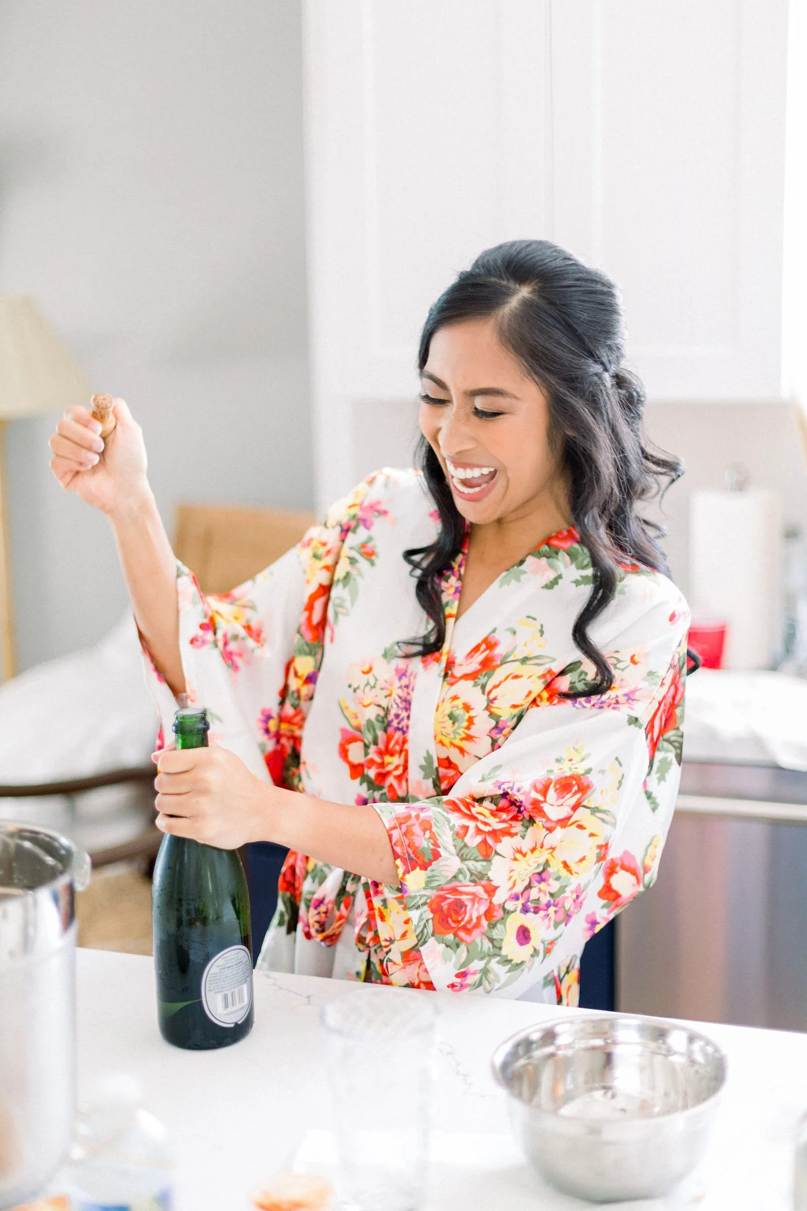 Bride in robe with soft glam makeup and styled hair holding a champagne glass while getting ready on wedding morning. 