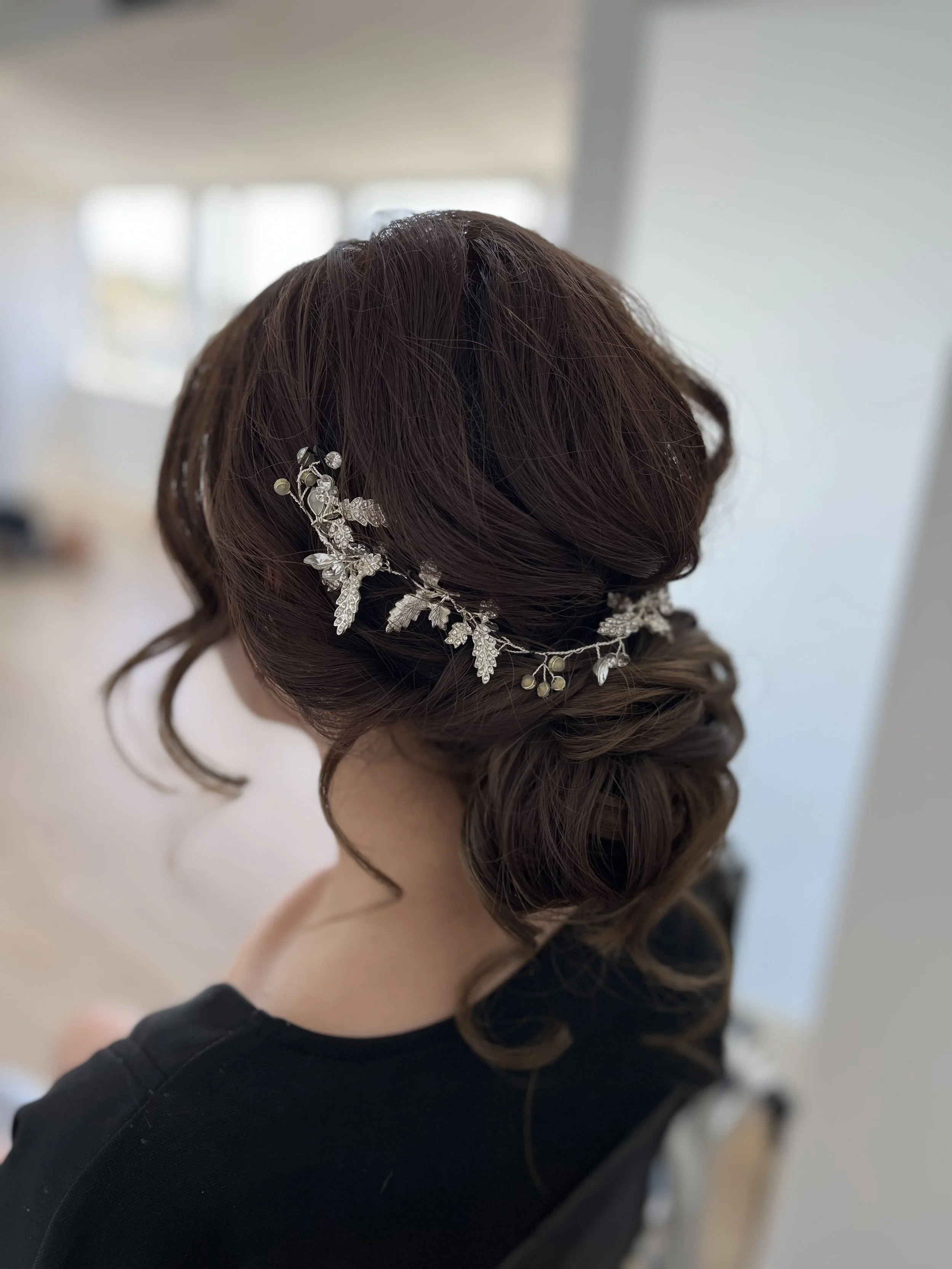 Low bridal bun with soft texture and delicate vine hair accessory, styled for a romantic wedding look. 