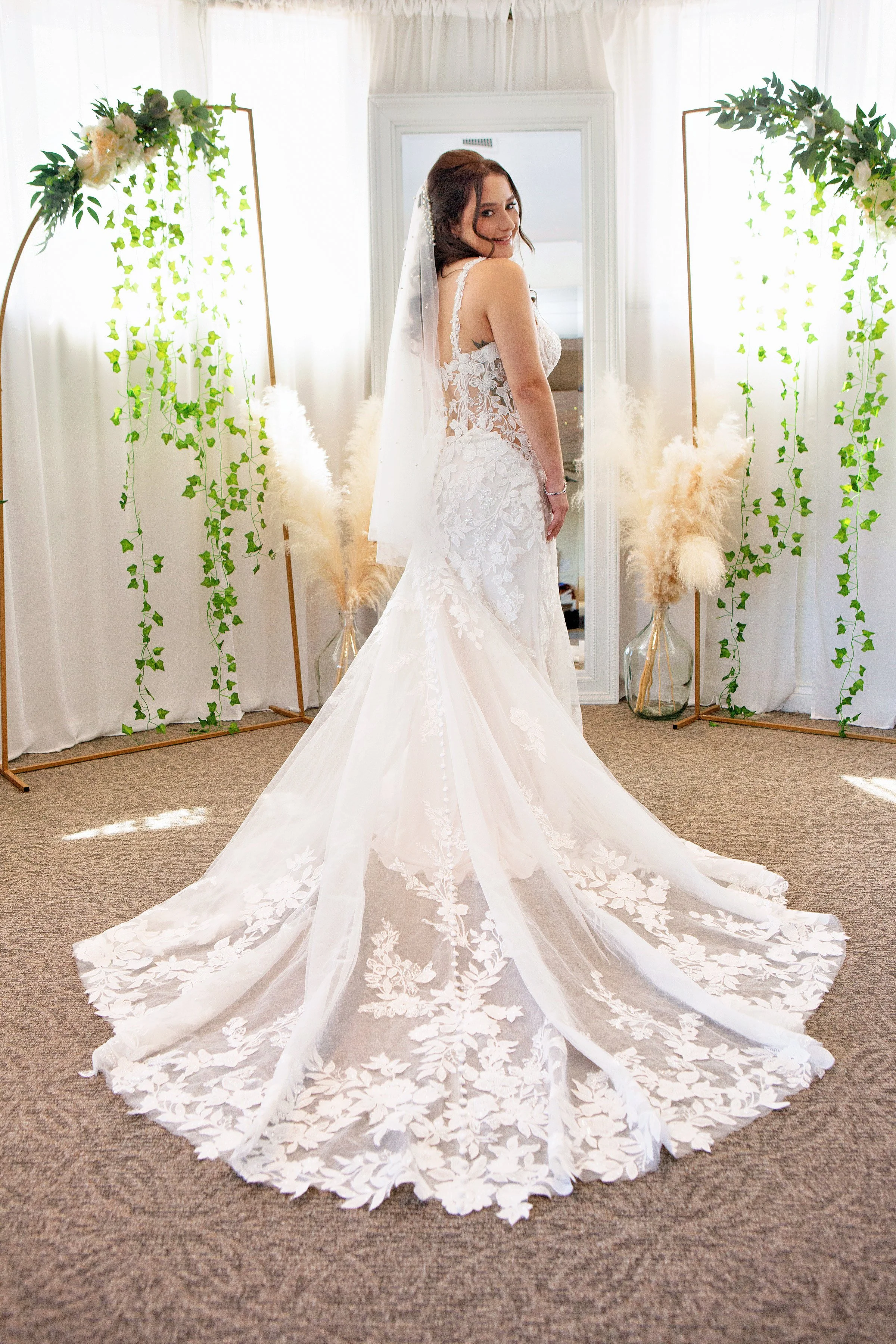 Bride in lace weddingf gown with veil stnading in front of a mirror, feautring soft glam bridal makeup and an elegant updo hairstyle.