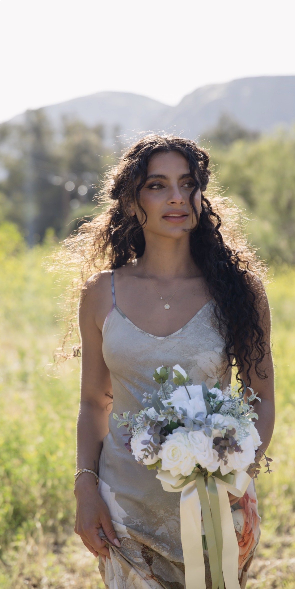 A woman with curly dark hair standing outdoors in a sunny field with hills in the background, holding a bouquet of white flowers, wearing a light-colored patterned dress, and looking to the side.