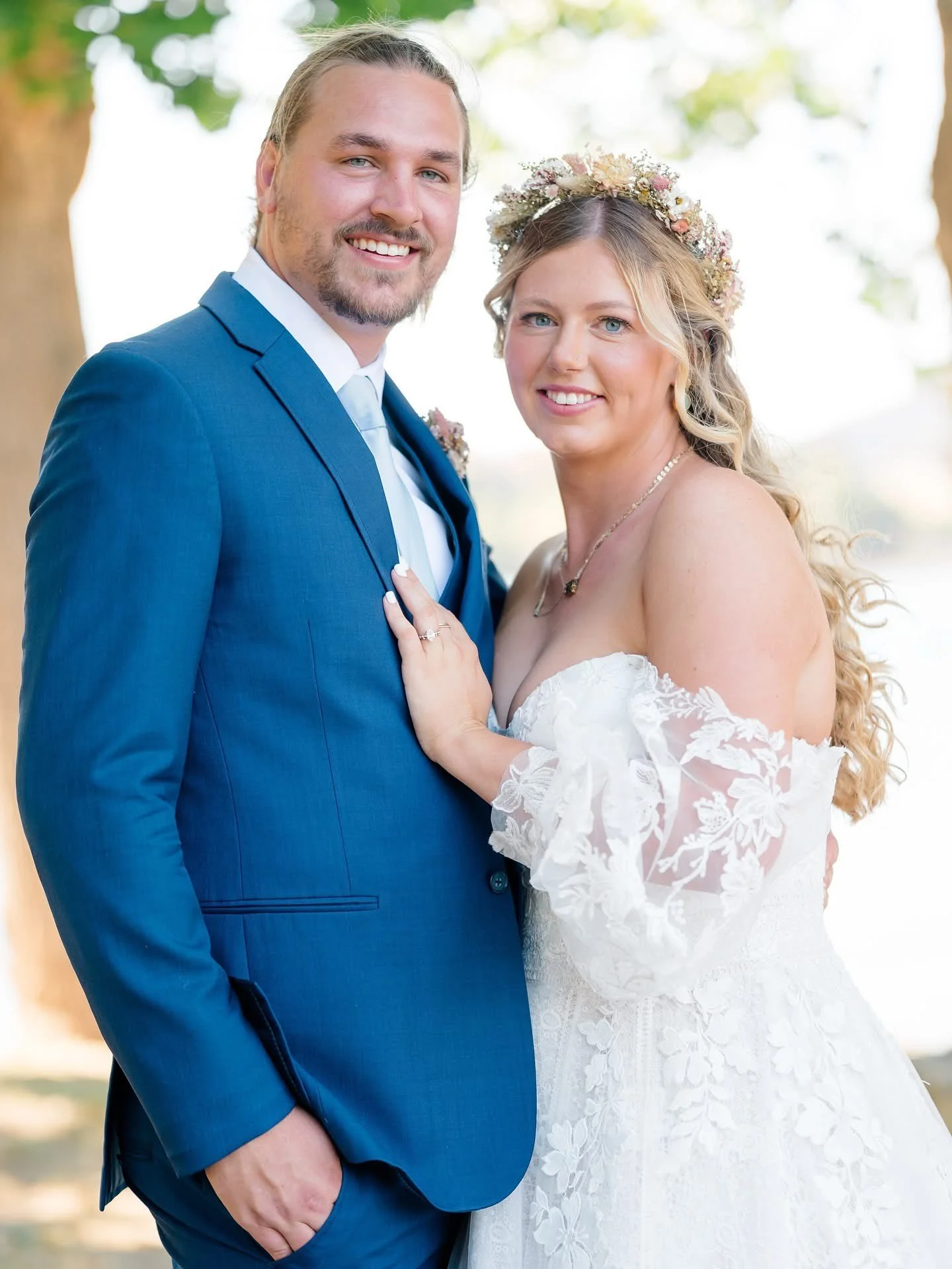 Bride smiling udner veil besides groom in blue suit during outdoor wedding ceremony.