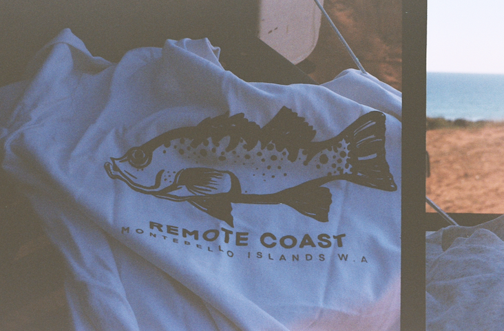 A white T-shirt with a fish graphic and the text 'REMOTE COAST' and 'MONTBELLO ISLANDS W.A.' printed on it, laying on a surface showing a view of the beach and ocean on a remote coastline..