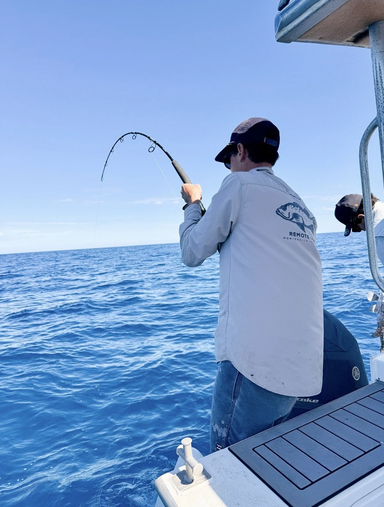 A person fishing off a boat in the ocean under a clear blue sky, wearing a remote coast white shirt, gray pants, and a cap.