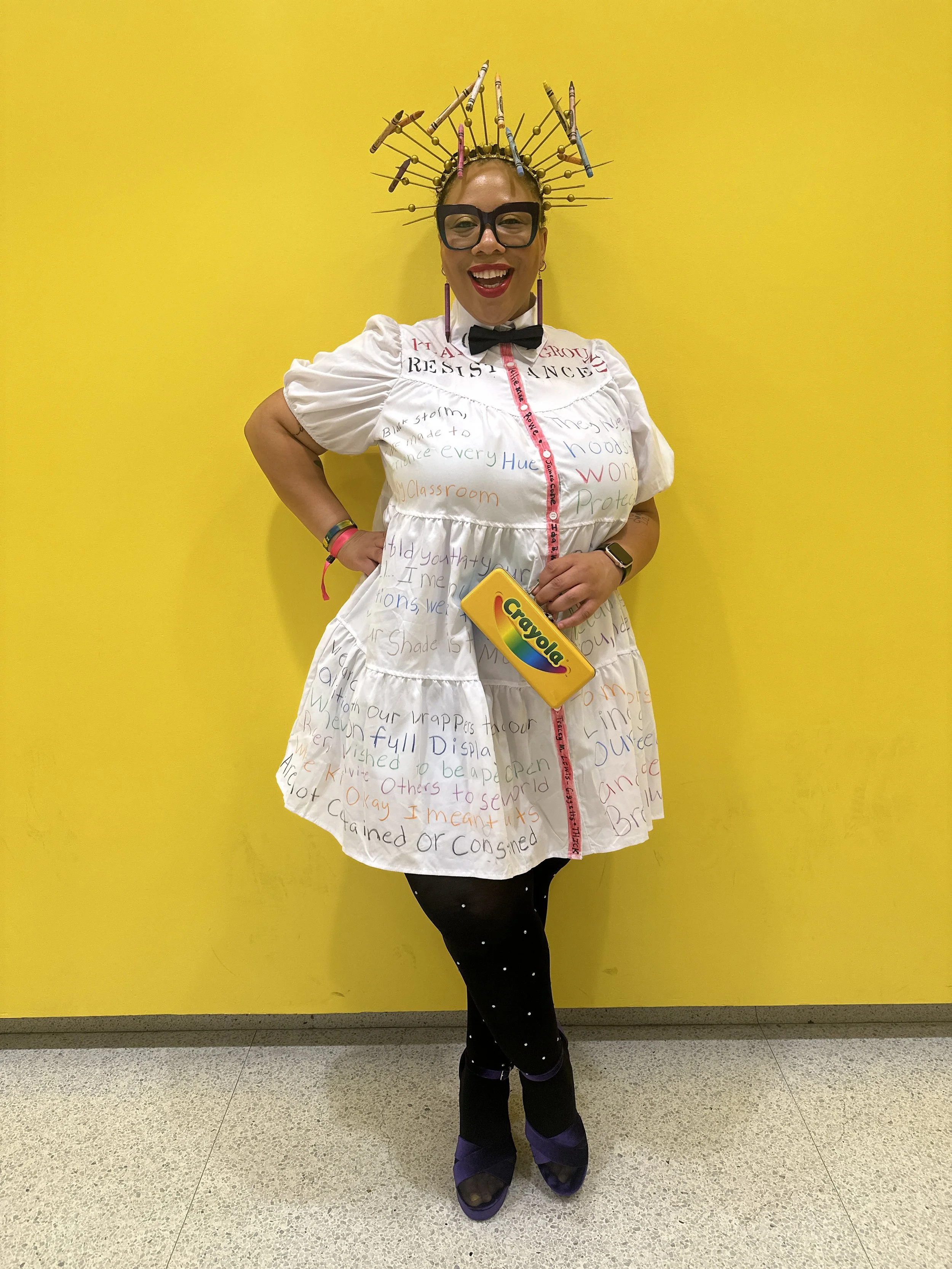 Woman dressed as a teacher, wearing glasses, a black bowtie, and a white dress with colorful words and phrases written on it, stands against a yellow wall. She is wearing a crown made of pencils, holding Crayola crayons, and smiling cheerfully.
