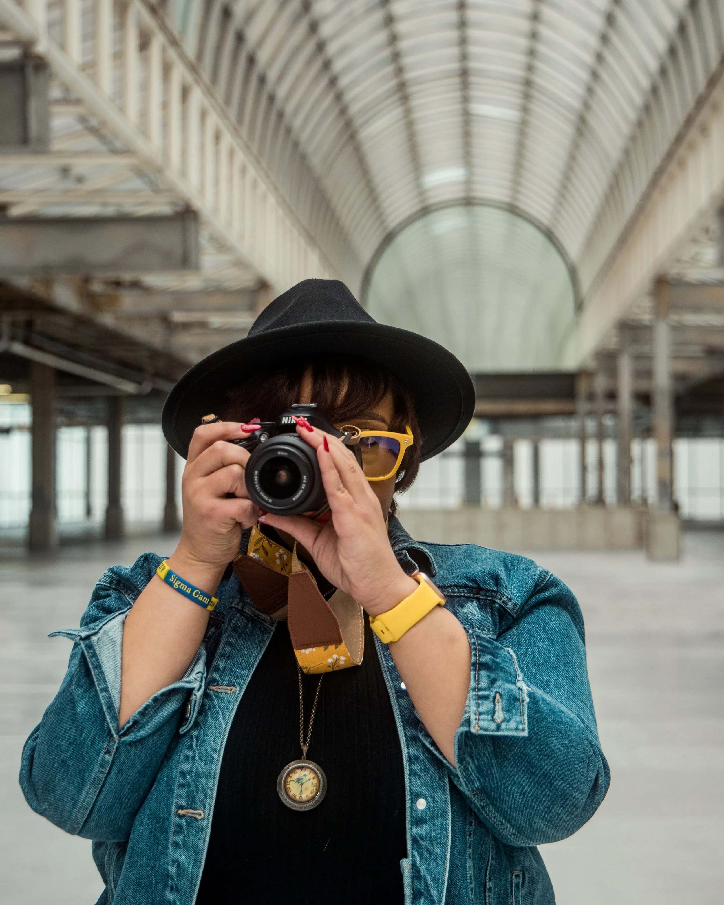 A woman in a denim jacket, black hat, and yellow glasses taking a photo with a camera inside a large glass and metal train station.