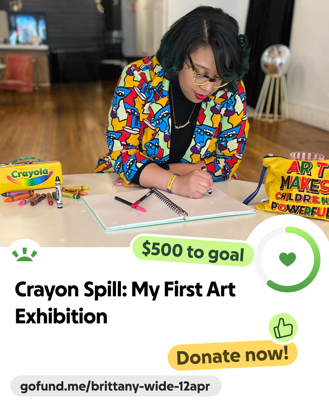 A woman with short black hair and colorful hair tips wearing glasses, a multicolored jacket with a cartoon face pattern, and a gold necklace, is sitting at a table drawing with markers. On the table are crayons, markers, a sketchbook, and a yellow bag with colorful text related to children's art. Background features a wooden floor, furniture, and a television.