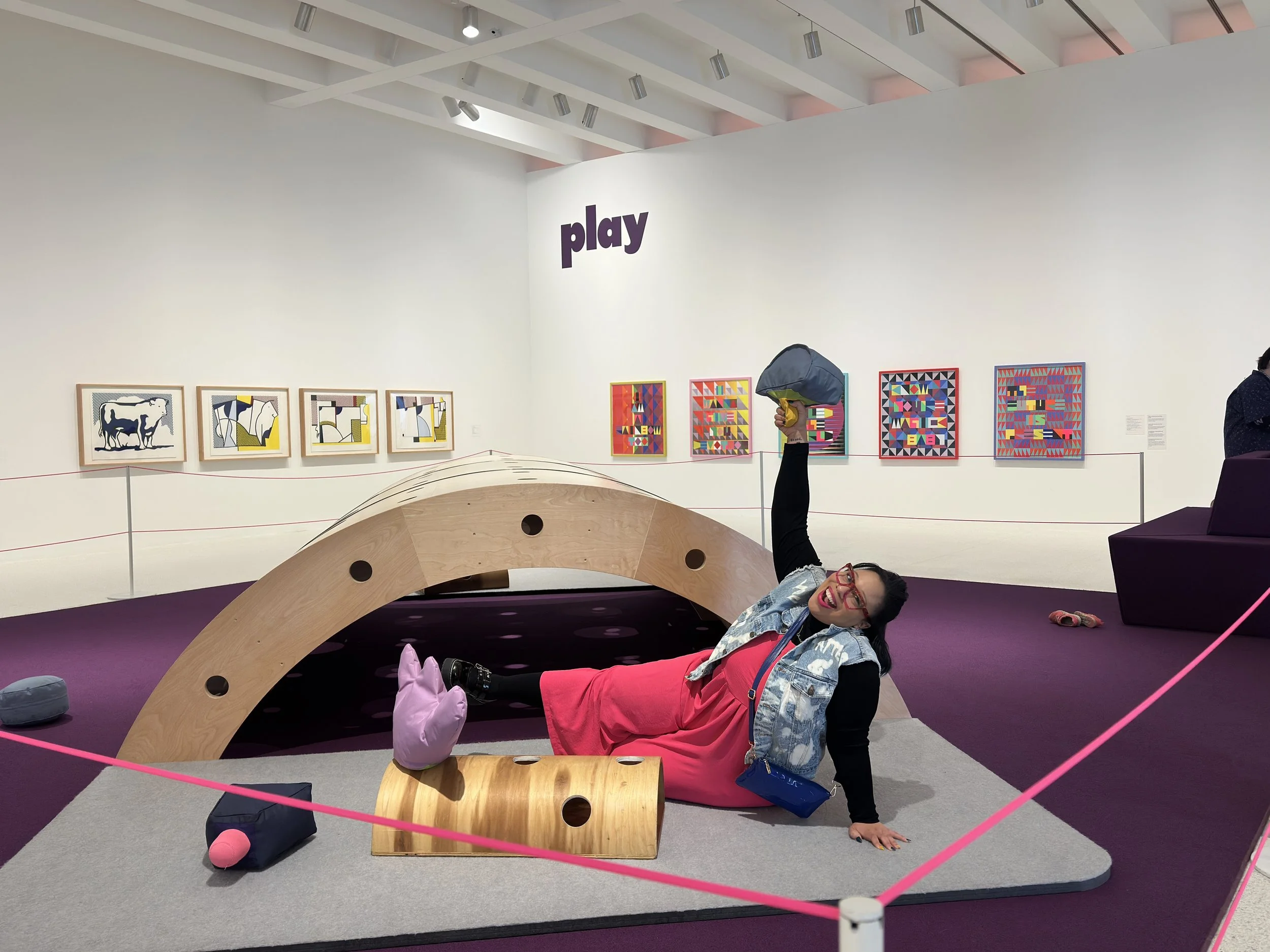 A woman with red glasses and a bright pink dress is lying on the floor of an art gallery, smiling and making a peace sign. She is under a wooden climbing structure and is holding a gray backpack in one hand. The gallery has colorful abstract paintings on the white walls, with a purple carpet and a pink cord fence around the display area.