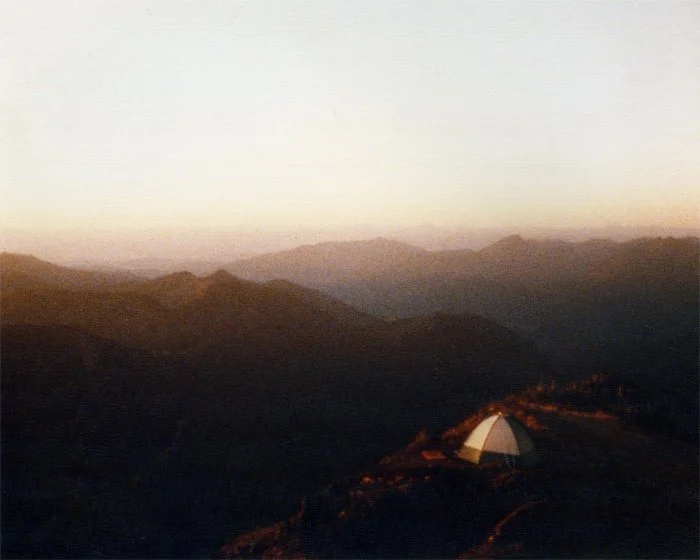 A lone tent illuminated at dusk on a high mountain ridge, with layered peaks receding into a hazy golden horizon