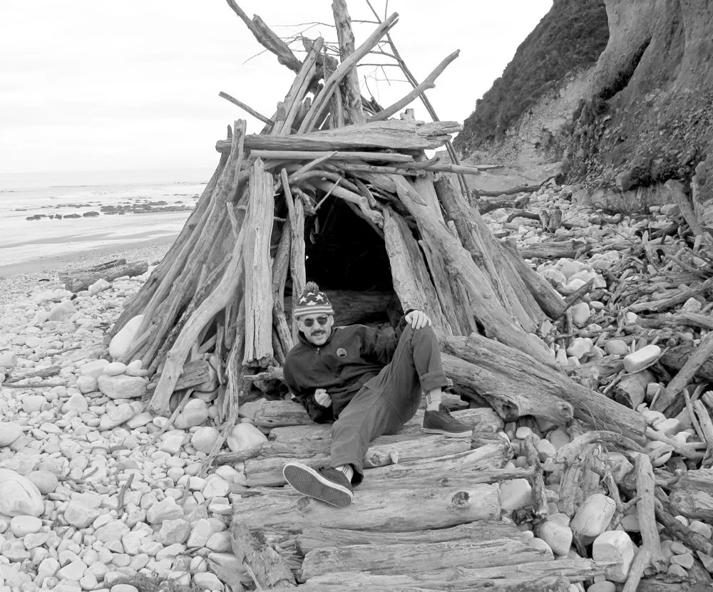 Una persona con gafas, barba y gorra de temática patriótica, recostada en la entrada de una estructura de madera construida con troncos en una playa con piedras, junto a un acantilado.