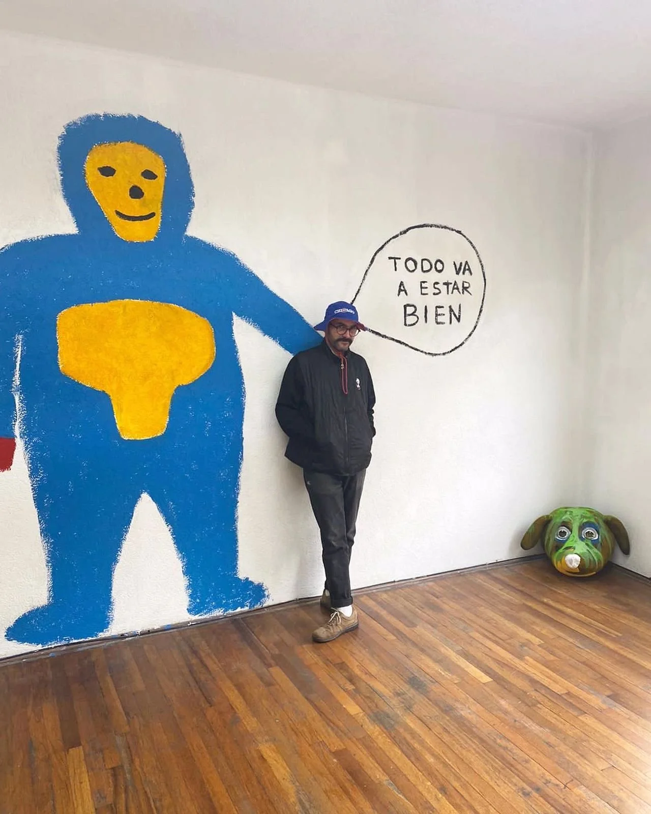 Hombre de pie junto a mural con dibujo de figura azul y amarilla, con globo de diálogo que dice 'Todo va a estar bien'. Está en una habitación con suelo de madera y una escultura de perro en una esquina.