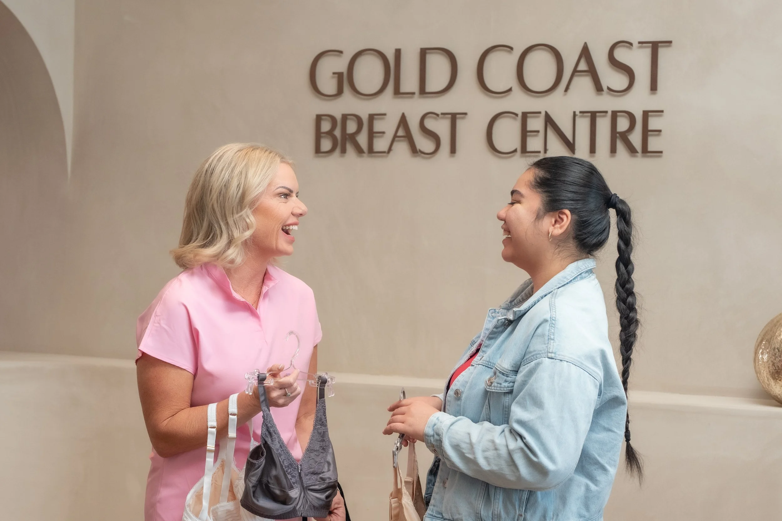 Two women, one in pink scrubs and the other in a denim jacket, are smiling and talking in front of a wall that reads 'Gold Coast Breast Centre.'