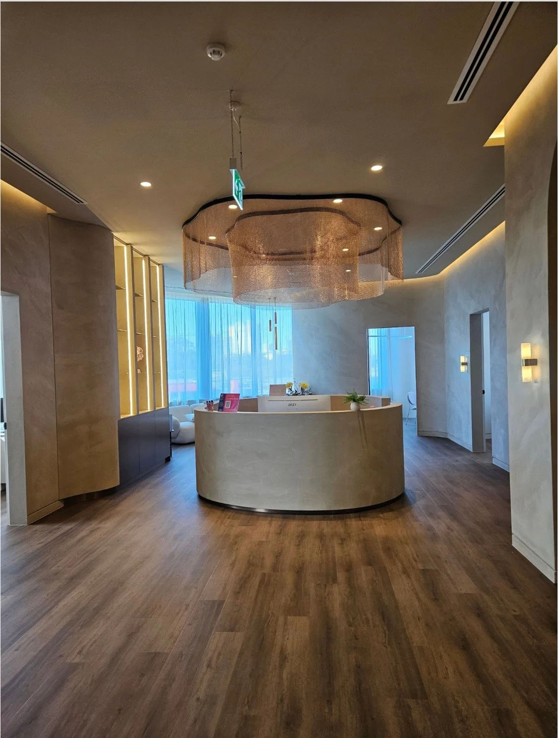 Modern hotel lobby with a curved reception desk, wooden flooring, a large window with sheer curtains, and contemporary lighting fixtures.