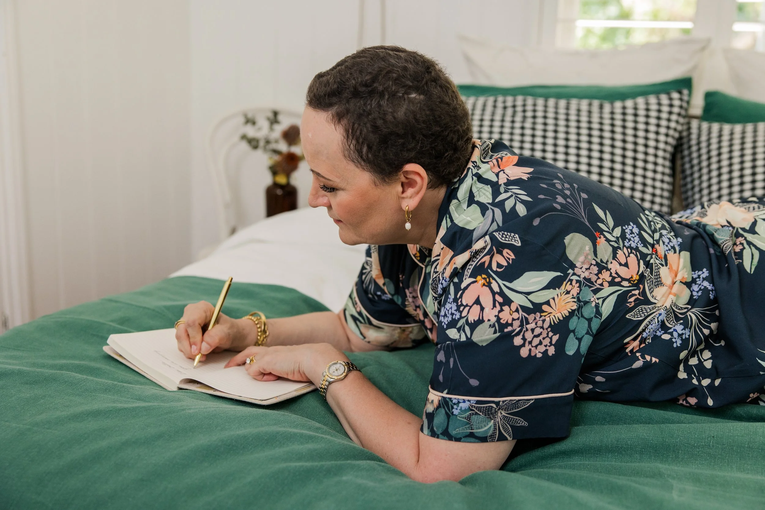Woman with short curly hair wearing a floral blouse, writing in a notebook on a green bedspread in a bright room.